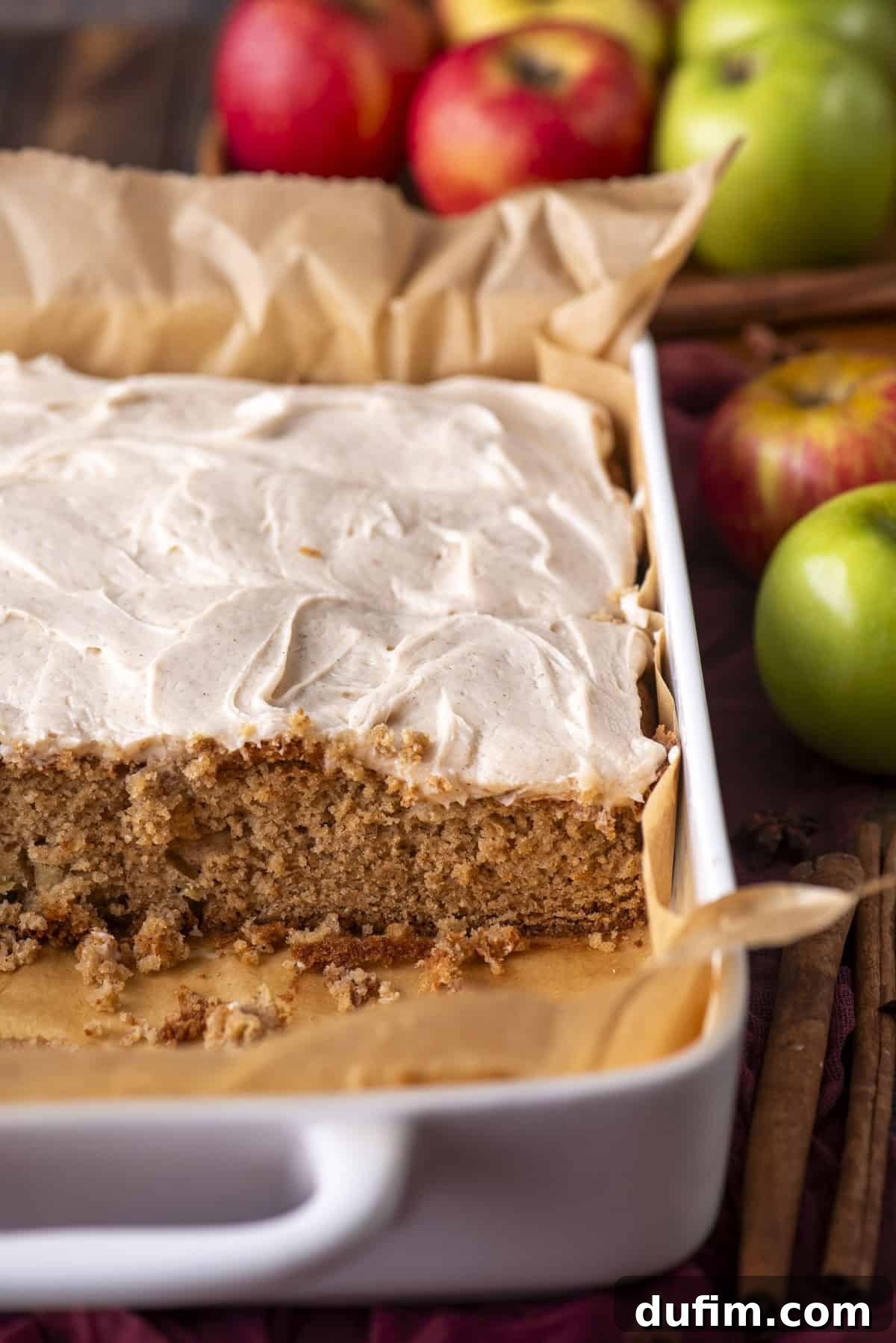 A white baking dish with a rectangular apple cake topped with creamy frosting, partially sliced. Whole red and green apples and cinnamon sticks are in the background. The cake sits on parchment paper.