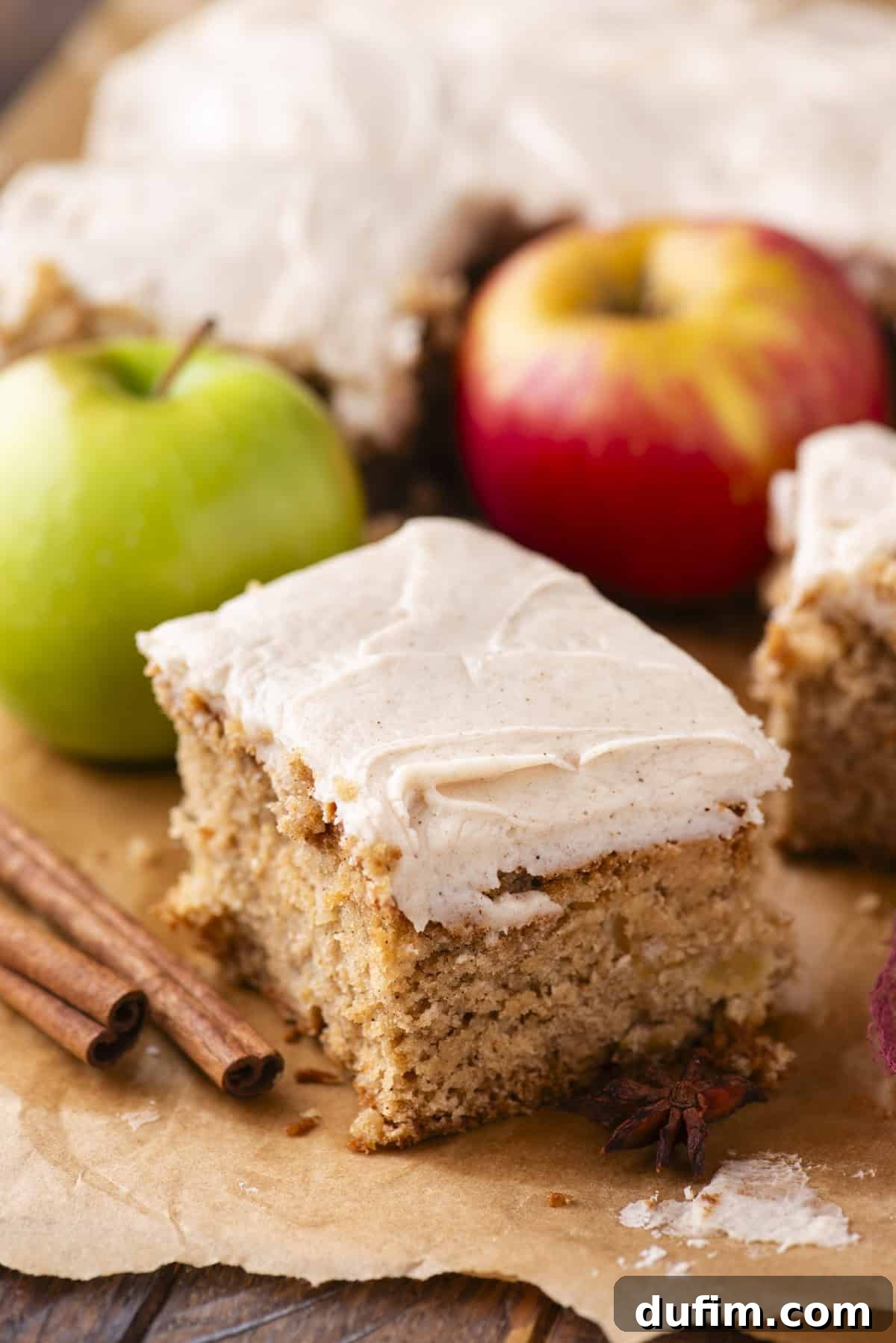 A slice of spiced apple cake with creamy frosting sits on parchment paper, surrounded by cinnamon sticks, a green apple, a red apple, and more cake in the background.