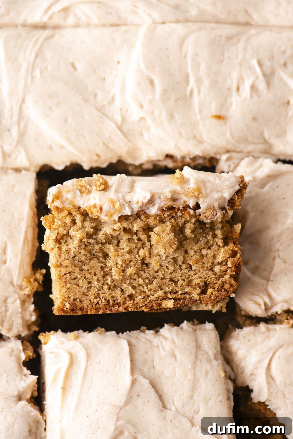 A close-up of a slice of moist, spiced cake with creamy frosting, lifted slightly above a tray of similar frosted cake pieces. The cake appears soft and dense with a thick layer of frosting on top.