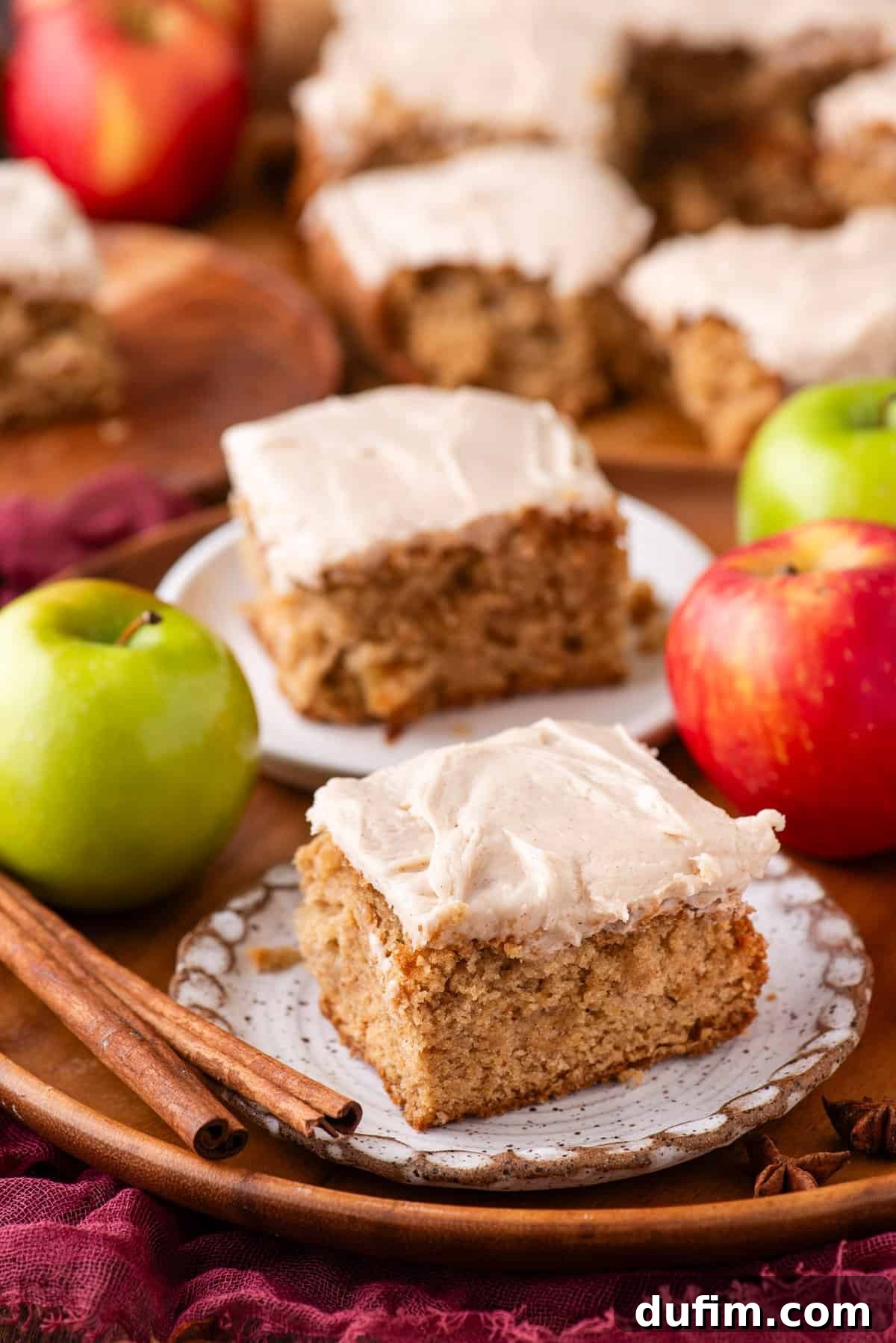 A close-up of frosted apple spice cake squares on plates, surrounded by fresh red and green apples and cinnamon sticks on a wooden tray. More cake pieces are visible in the background.