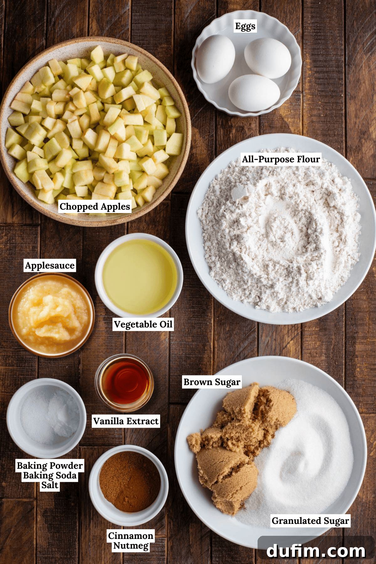 Ingredients for an Apple Bundt Cake are arranged on a wooden table: chopped apples, eggs, all-purpose flour, applesauce, vegetable oil, brown sugar, granulated sugar, vanilla extract, cinnamon, nutmeg, baking powder, baking soda, and salt.