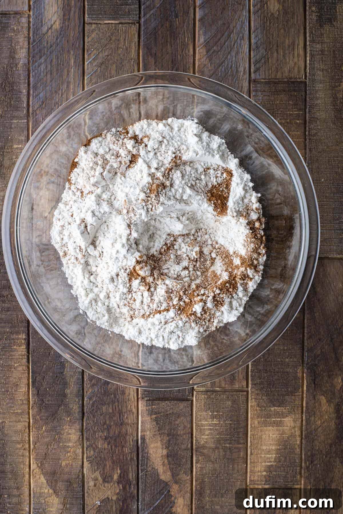A glass bowl filled with flour and cinnamon sits on a dark wooden surface. The ingredients are unmixed, with visible streaks of brown cinnamon on top of the white flour.
