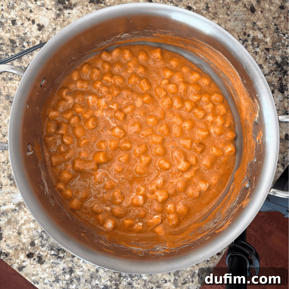 A stainless steel pot filled with mini marshmallows melting into a thick, orange sauce sits on a speckled granite countertop. The mixture appears creamy and smooth, partially coating the sides of the pot.
