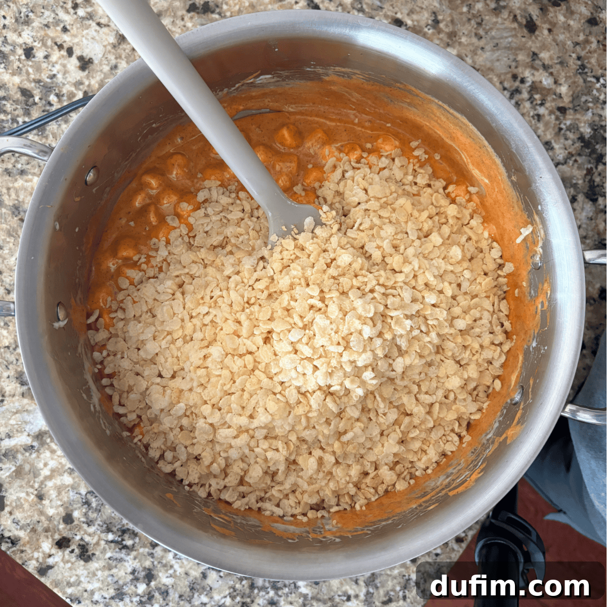 A saucepan on a granite countertop contains a mixture of melted orange marshmallow or caramel with a layer of rice cereal on top and a white spatula resting inside.