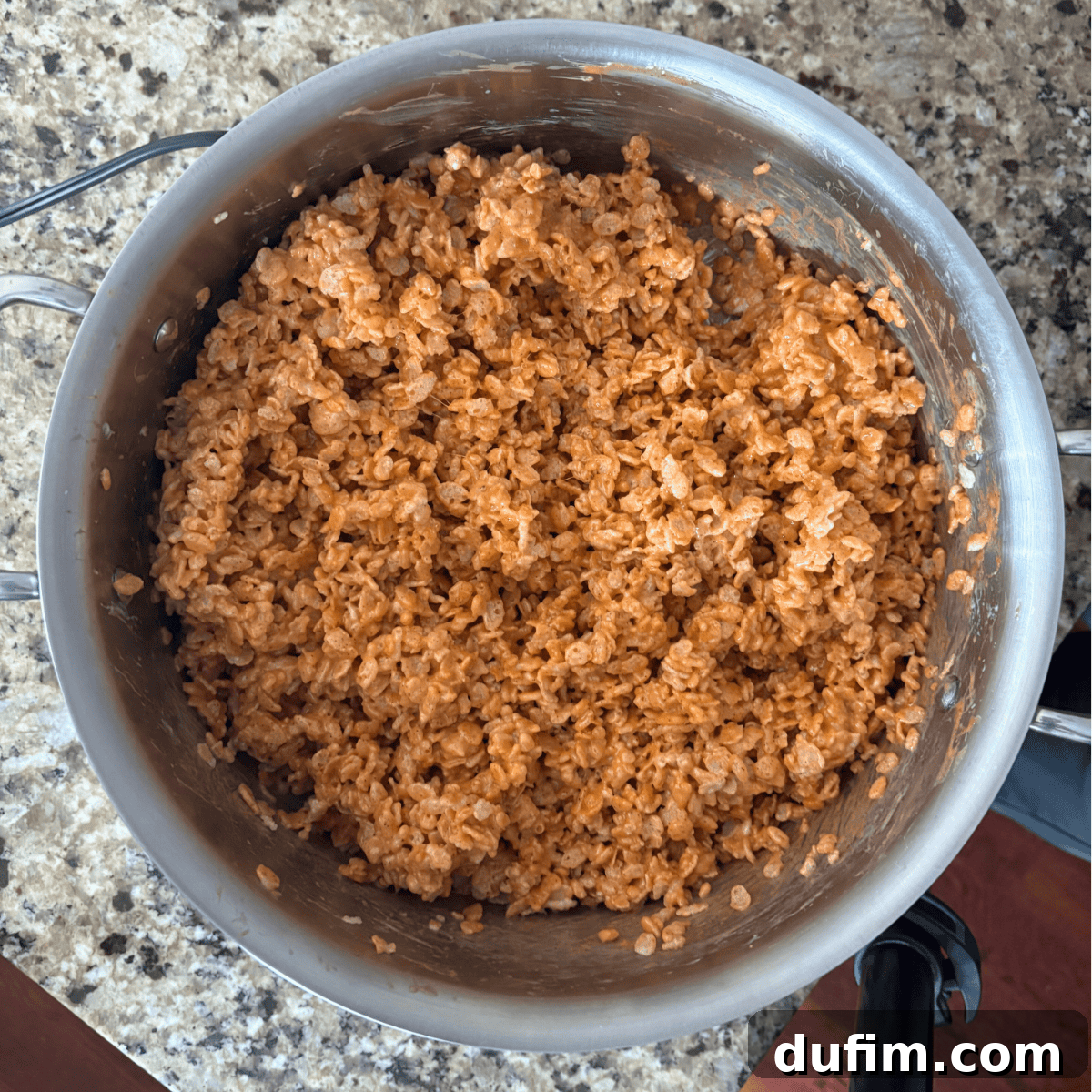 A stainless steel pot on a granite countertop filled with crispy rice cereal mixed with melted chocolate and possibly peanut butter or another brown ingredient.