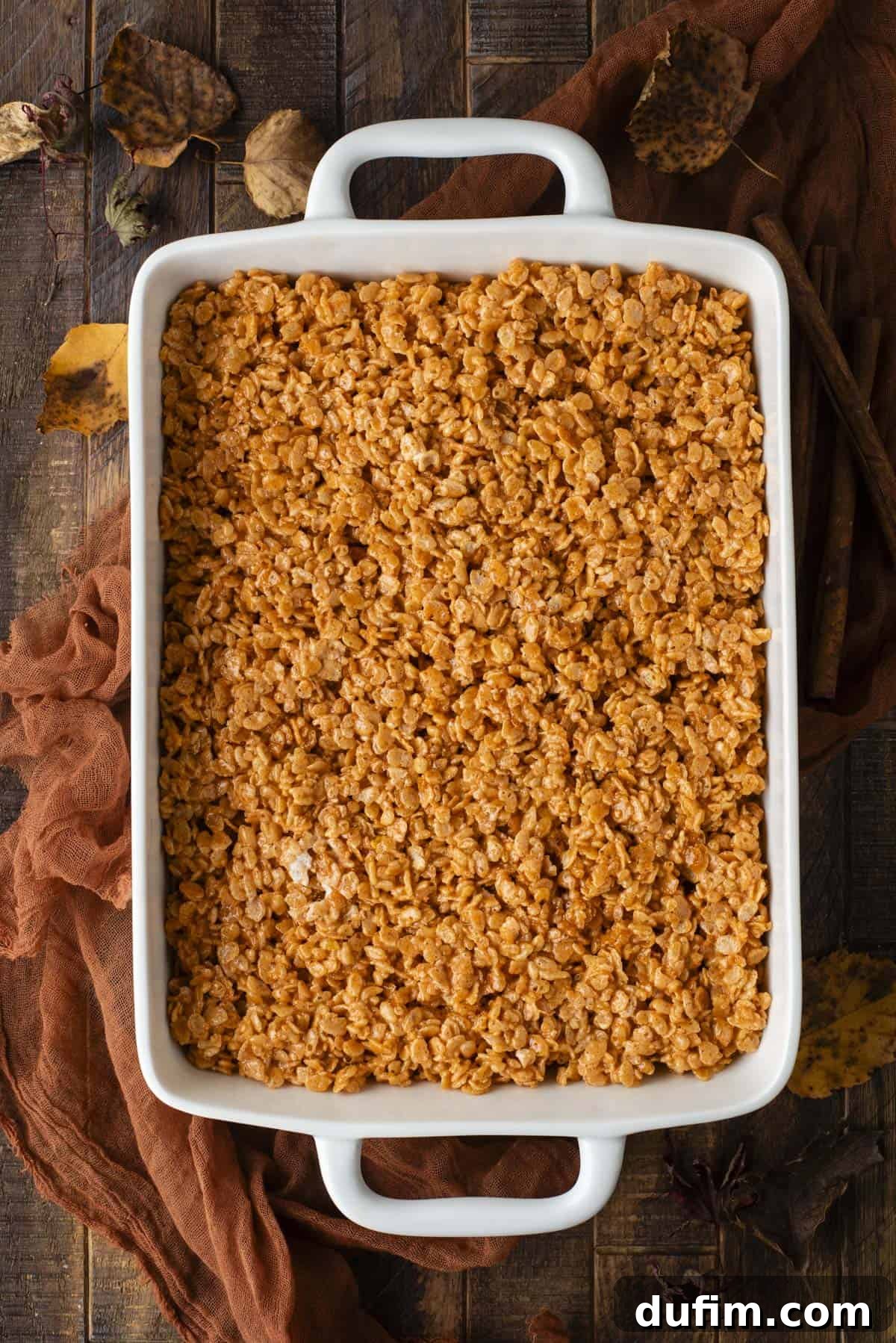 A white rectangular baking dish filled with golden-brown Rice Krispies treats sits on a rustic wooden table, surrounded by brown cloth and autumn leaves.