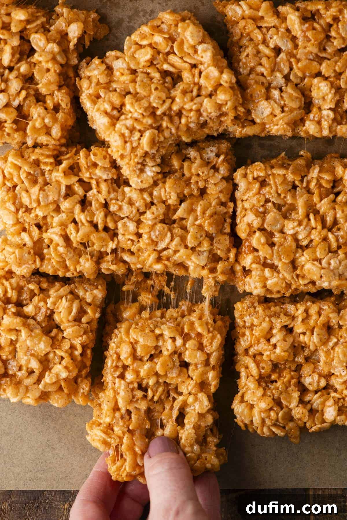 A hand is pulling apart a square of sticky, golden rice crispy treat from a batch of similar squares on a brown surface. The treats have a glossy, marshmallow-coated appearance.