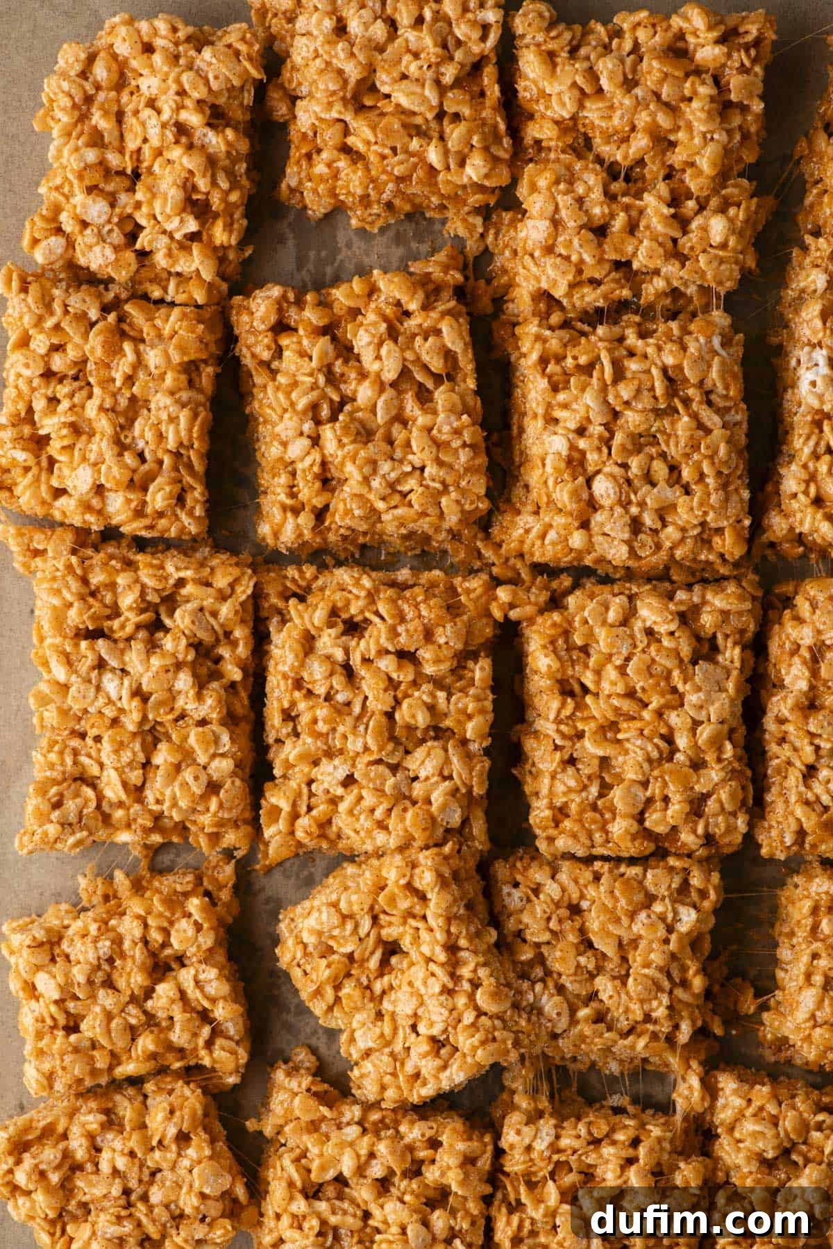 Close-up of several square Rice Krispies treats arranged in rows on parchment paper, showing their crispy, golden-brown texture and slightly sticky appearance.