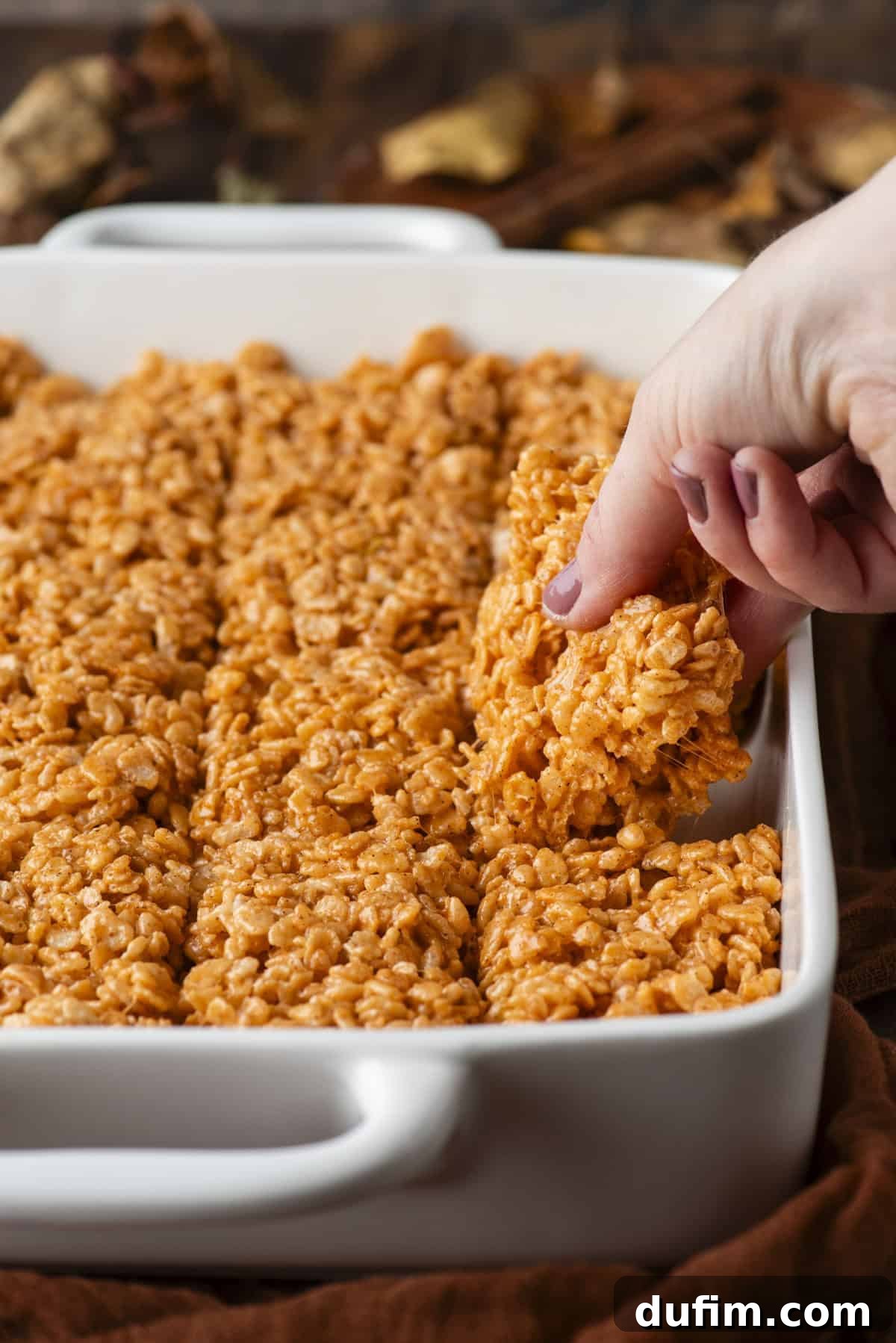 A hand lifts a pumpkin-colored rice crispy treat from a white baking dish filled with neatly cut treats.