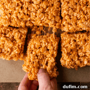 A hand lifts a gooey rice crispy treat from a batch of square-cut treats on parchment paper. The treats are golden brown with visible puffed rice cereal, stuck together with melted marshmallow.