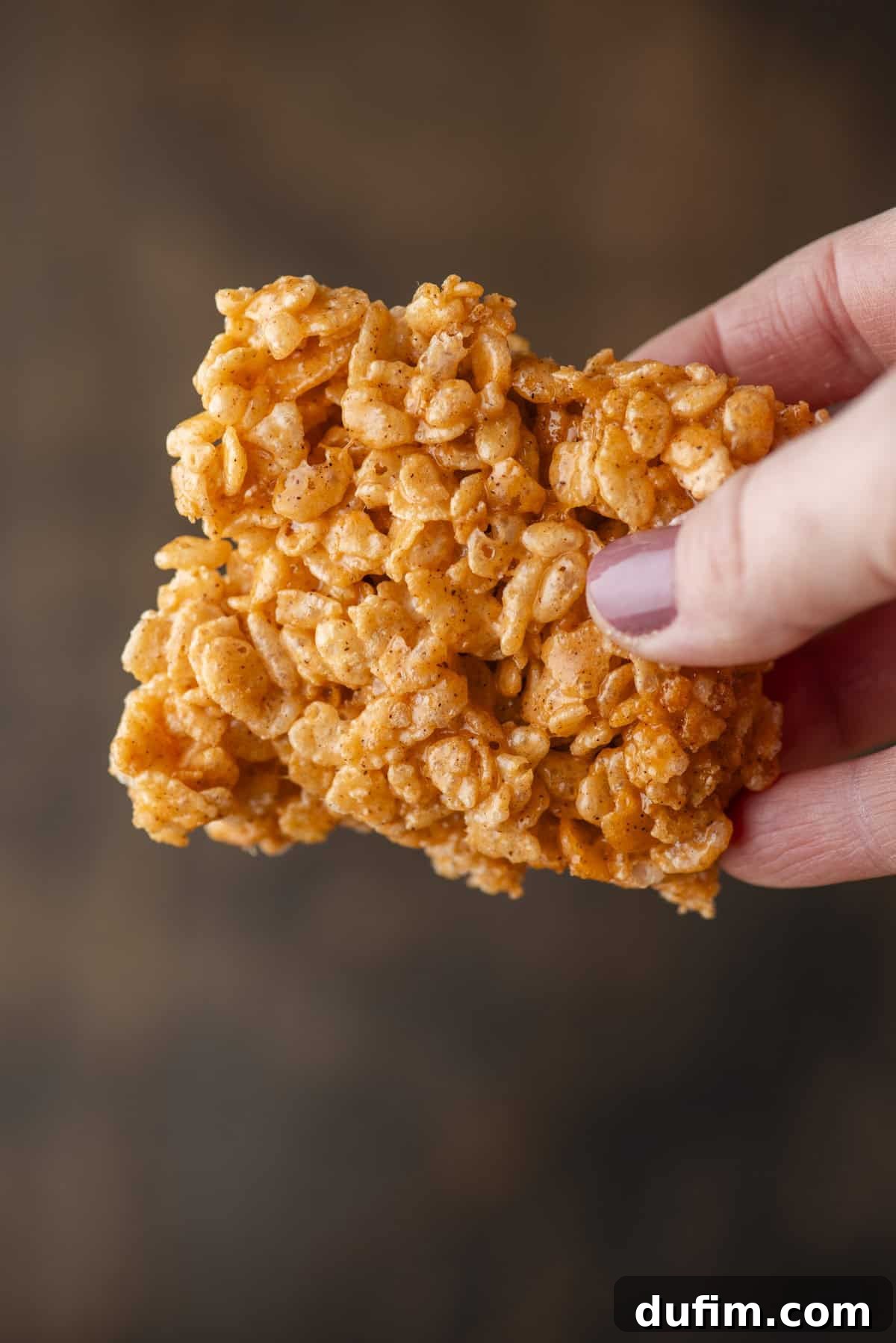 A hand with painted nails holds a single Rice Krispies treat made of puffed rice cereal and marshmallow against a blurred brown background.