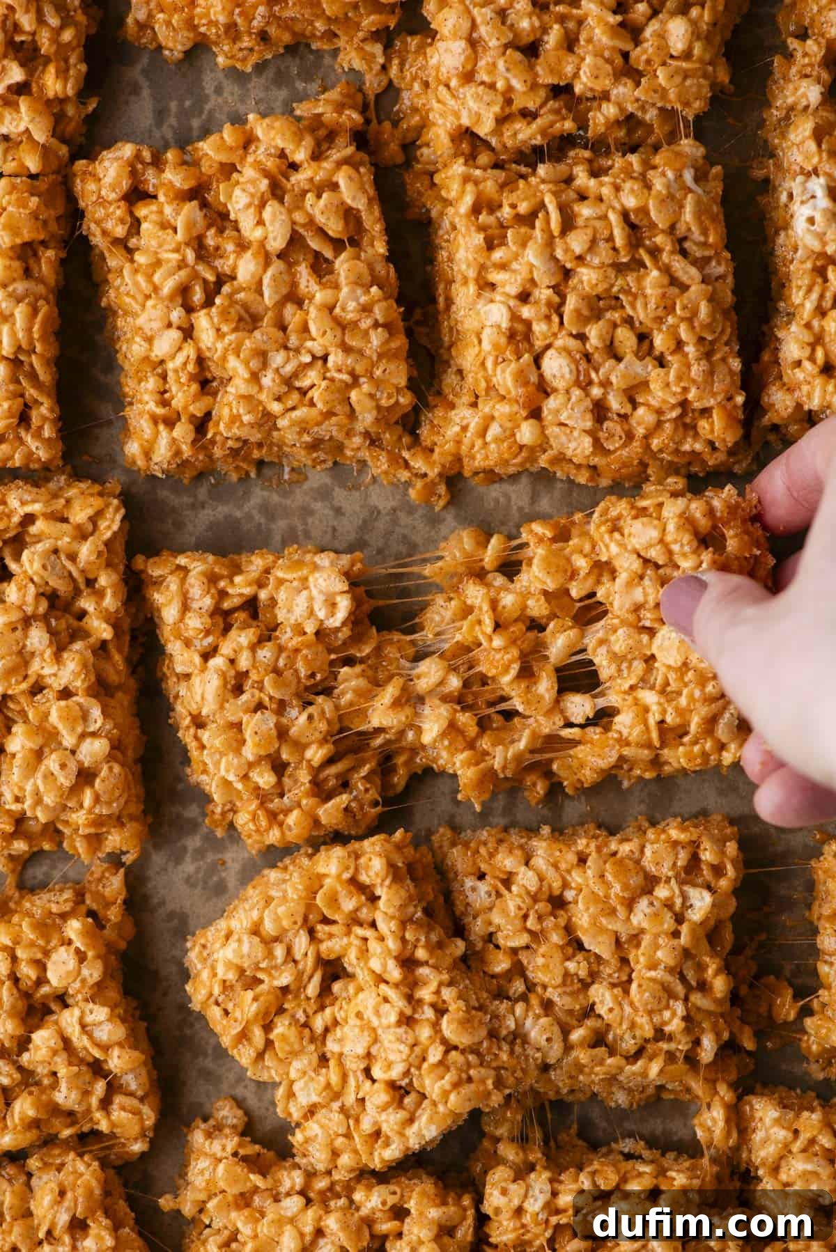 Hand pulling apart a square of golden brown Rice Krispies treat, showing gooey, sticky texture. Several other squares are arranged closely on parchment paper in the background.