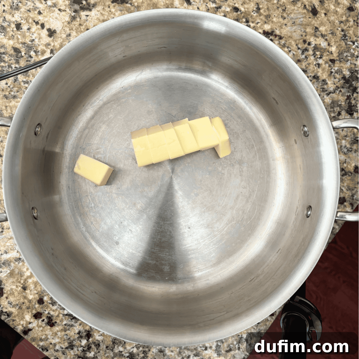 A stainless steel pot contains several small cubes of butter arranged in a row on its bottom, sitting on a speckled granite countertop.