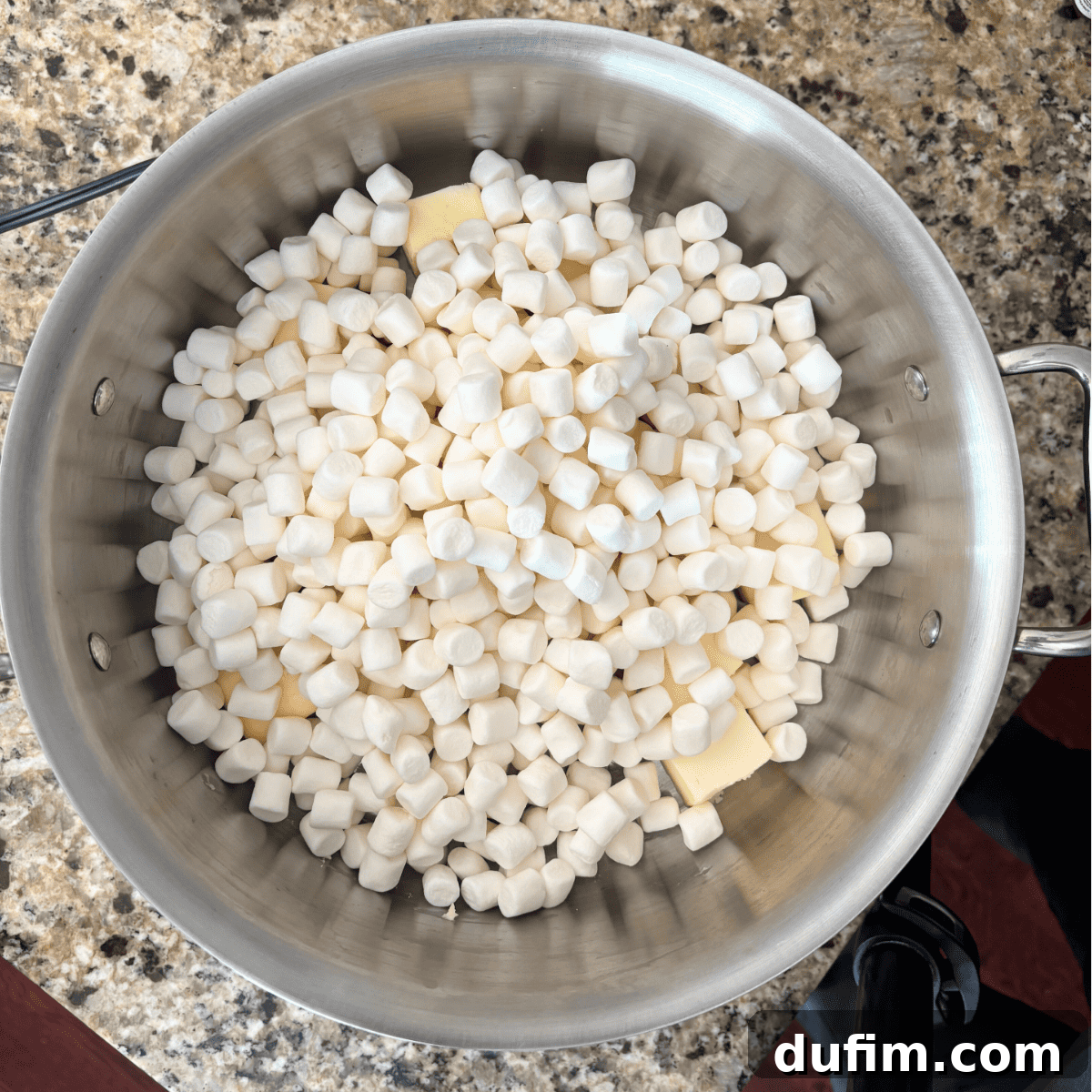 A large silver pot on a granite countertop filled with mini marshmallows and pieces of butter, ready to be melted together.