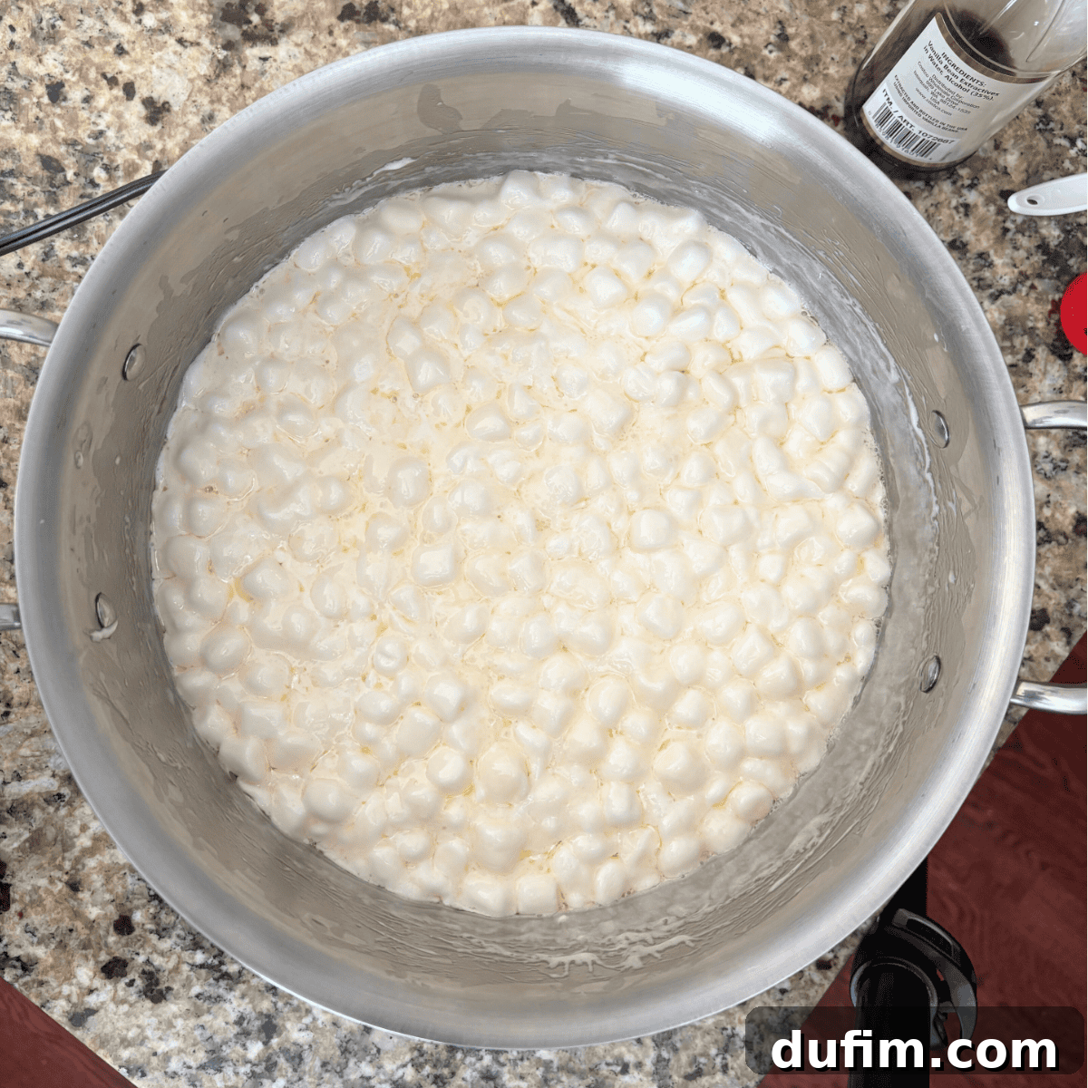 A large silver pot on a granite countertop containing melted mini marshmallows. A bottle of vanilla extract is visible nearby.