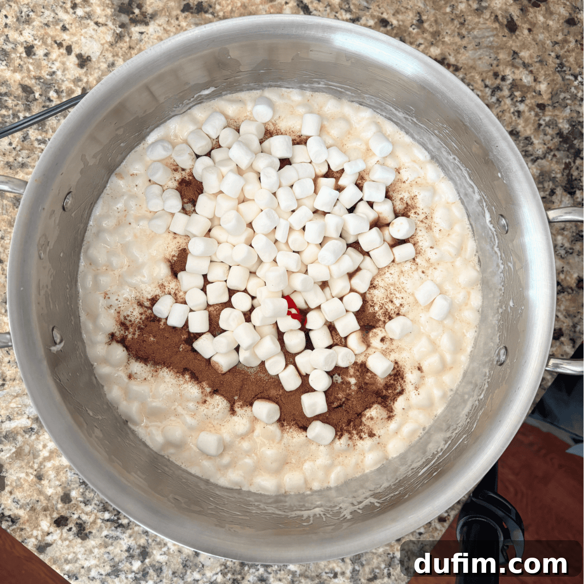 A pot on a granite countertop filled with melted marshmallows, mini marshmallows, cinnamon, and a drop of red food coloring, ready to be mixed.