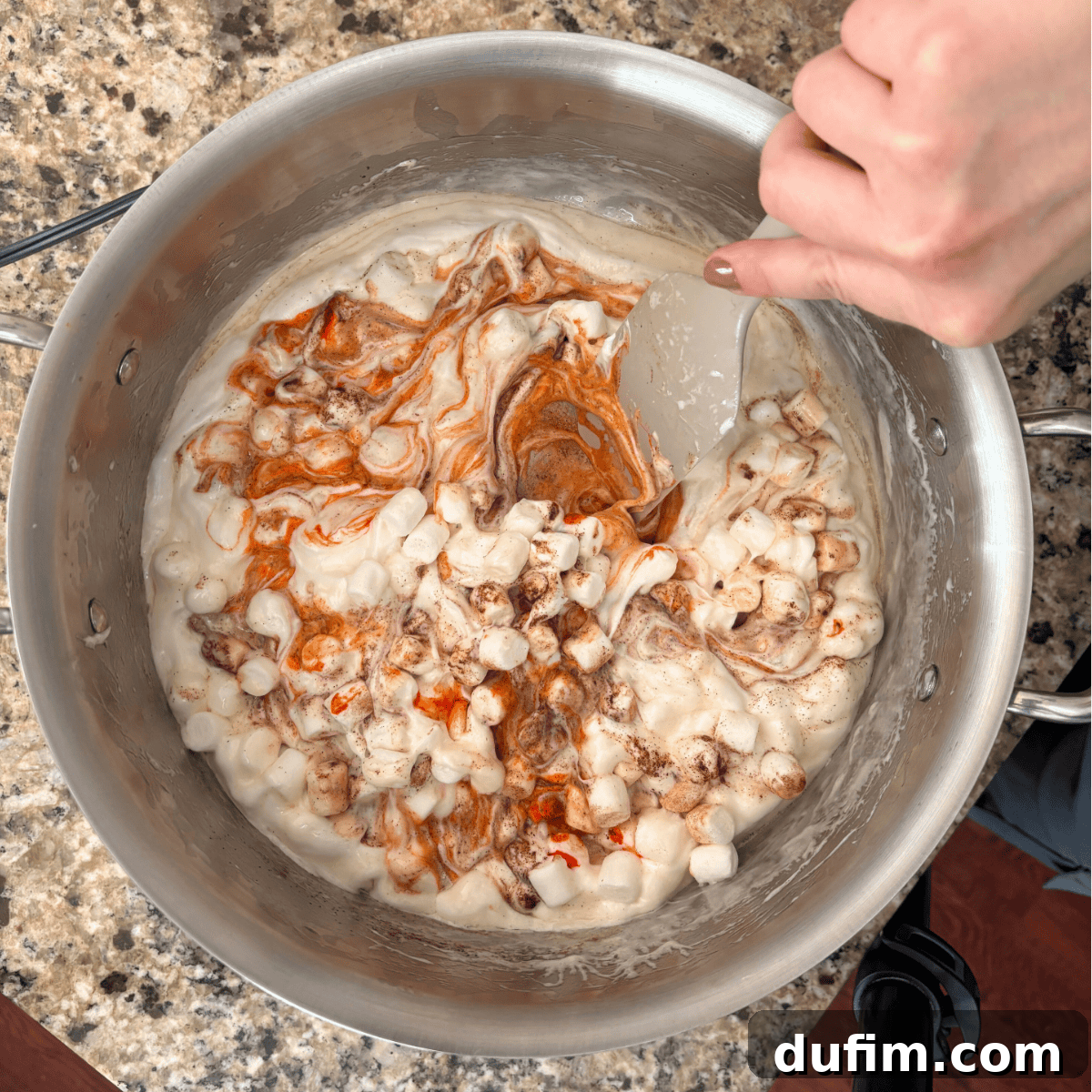 A hand holding a spatula stirs mini marshmallows with melted butter and swirls of orange coloring in a large metal pot on a granite countertop.