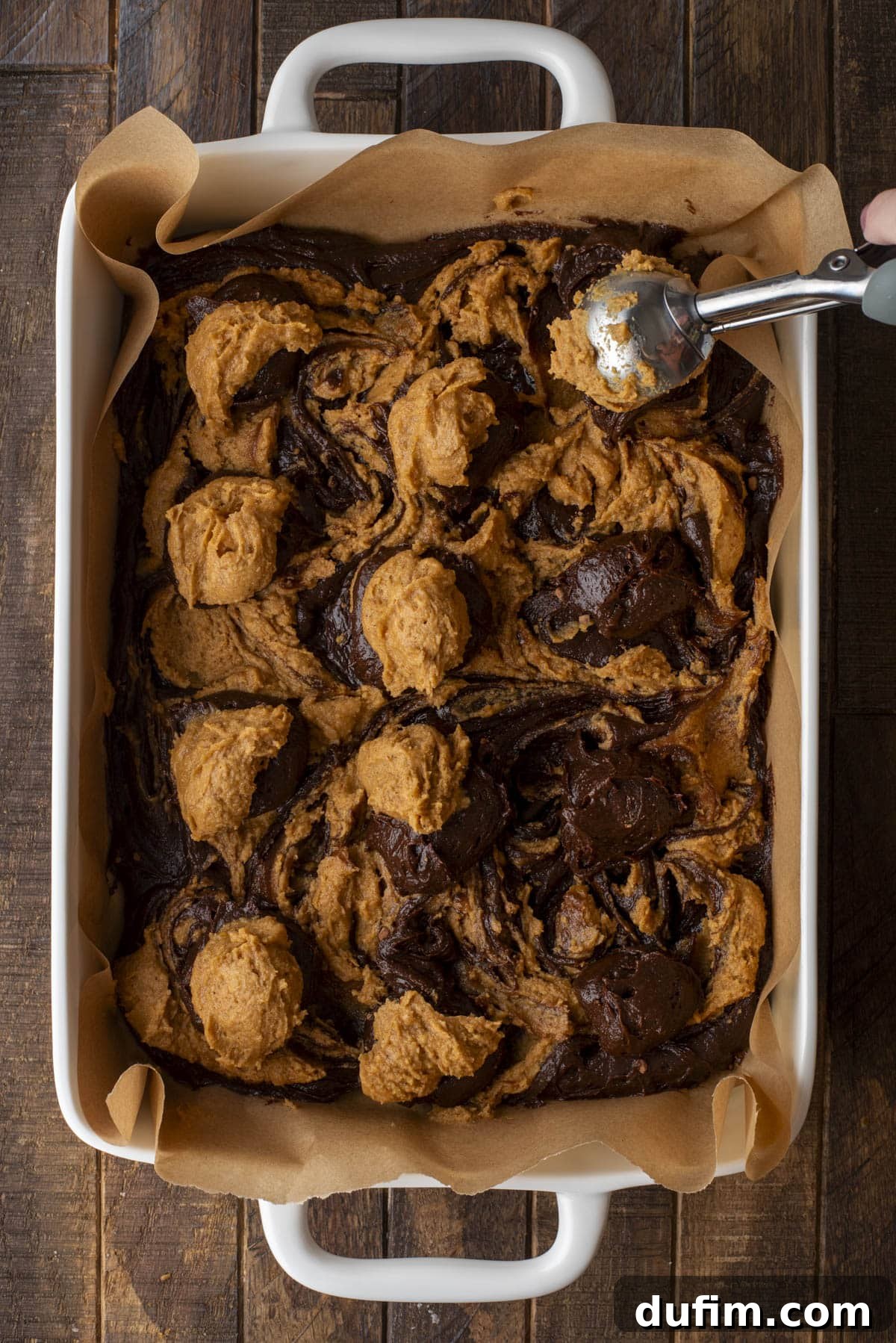 Pumpkin Ripple Fudge Brownies 11 A baking dish lined with parchment paper contains swirled dollops of chocolate and pumpkin brownie batter. A hand is using a cookie scoop to add more batter, preparing for the next swirl.