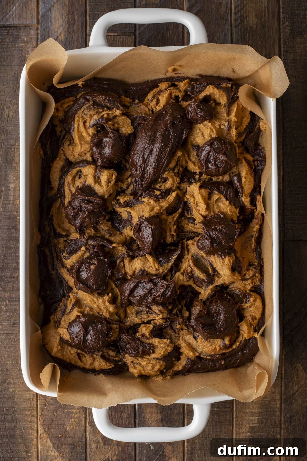 Pumpkin Ripple Fudge Brownies 12 A white baking dish lined with parchment paper holds a freshly baked dessert with dark chocolate and golden orange pumpkin swirls, giving a beautifully marbled appearance. The dish is placed on a rustic wooden surface, showing the rich texture.
