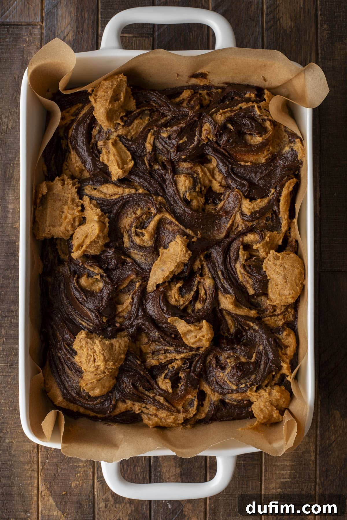 Pumpkin Ripple Fudge Brownies 13 A rectangular baking dish lined with parchment paper holds freshly baked brownies swirled with a lighter, golden brown pumpkin mixture on top. The dessert has a stunning marbled pattern and rests on a warm wooden surface.