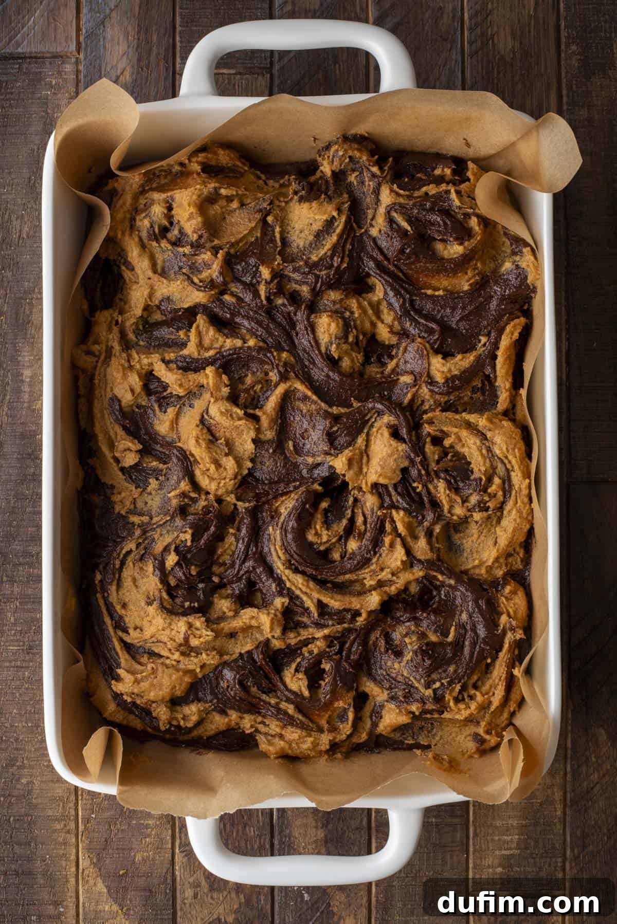 Pumpkin Ripple Fudge Brownies 14 A rectangular baking dish lined with parchment paper holds a batch of expertly swirled brownies, featuring a marbled mix of dark chocolate and lighter, caramel-colored pumpkin batter. The brownies appear freshly baked, rich, and enticingly textured.