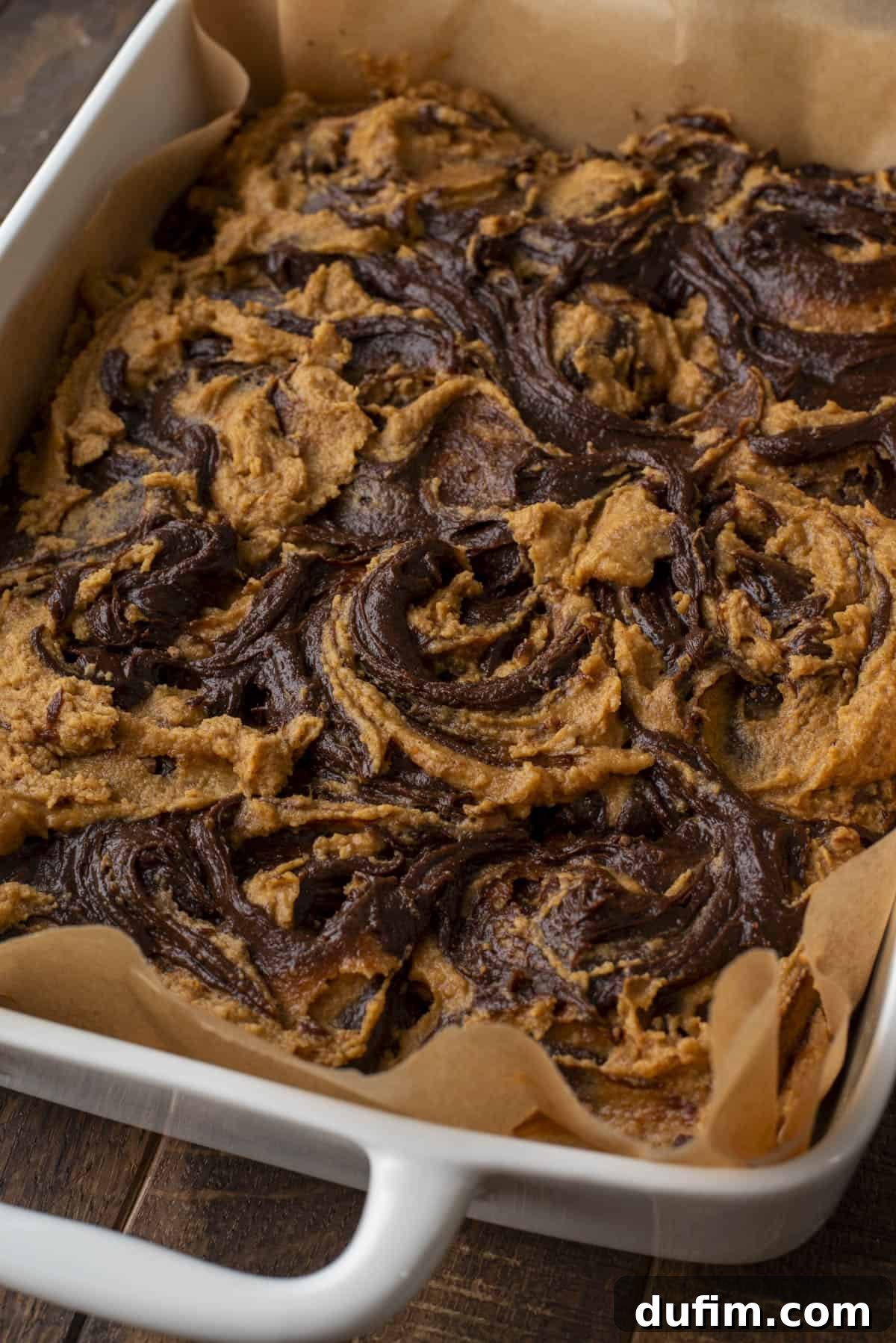 Pumpkin Ripple Fudge Brownies 15 A close-up of a square baking dish lined with parchment paper, filled with a dessert featuring beautifully swirled layers of dark chocolate and vibrant orange pumpkin. The dessert is freshly baked and slightly golden at the edges, hinting at its deliciousness.