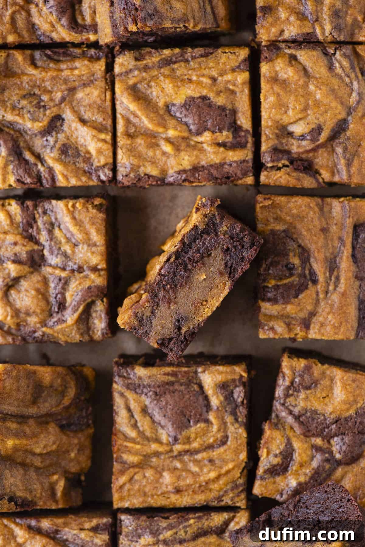 Pumpkin Ripple Fudge Brownies 18 A batch of fudgy brownies with a marbled pattern, featuring beautiful swirls of dark chocolate and golden orange pumpkin layers. One brownie is thoughtfully turned on its side, clearly showing the contrasting, delectable layers inside.