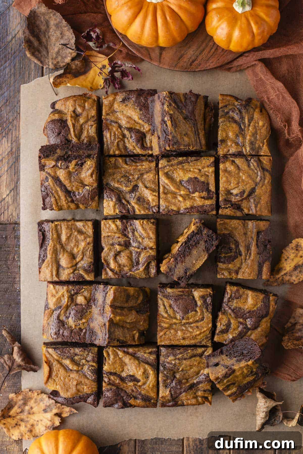 Pumpkin Ripple Fudge Brownies 6 Top-down view of pumpkin swirl brownies cut into neat squares on parchment paper, beautifully arranged and surrounded by scattered autumn leaves, mini pumpkins, and a brown cloth, set on a rustic wooden table. The marbled chocolate and orange pattern is prominent.