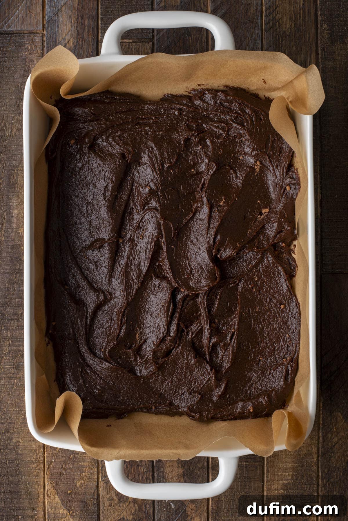 Pumpkin Ripple Fudge Brownies 7 A white baking dish lined with brown parchment paper filled with unbaked chocolate brownie batter, spread evenly on a dark wooden surface.