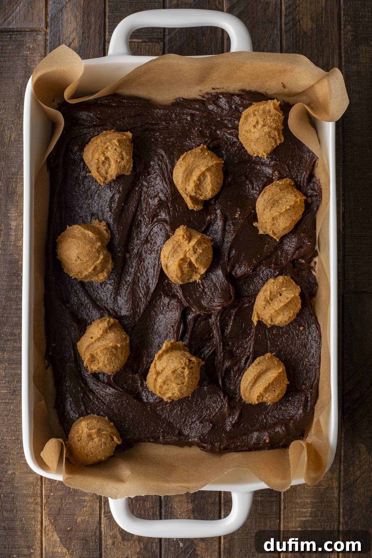 Pumpkin Ripple Fudge Brownies 8 A white baking dish lined with parchment paper contains chocolate brownie batter spread out, with several scoops of vibrant orange pumpkin mixture evenly placed on top, ready for swirling.