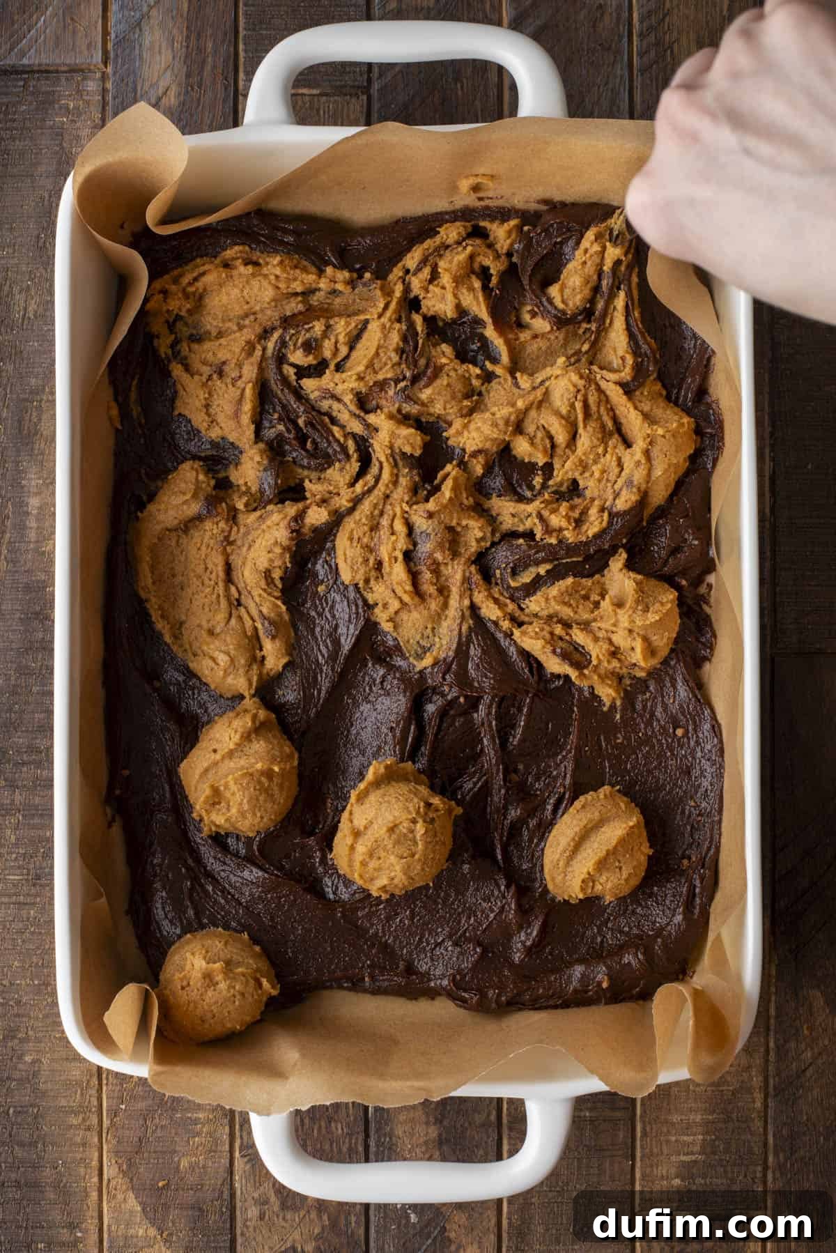 Pumpkin Ripple Fudge Brownies 9 A hand uses a knife to gently swirl dollops of orange pumpkin batter into chocolate brownie batter in a parchment-lined baking dish, creating a marbled effect. Some distinct scoops of pumpkin batter are still visible.