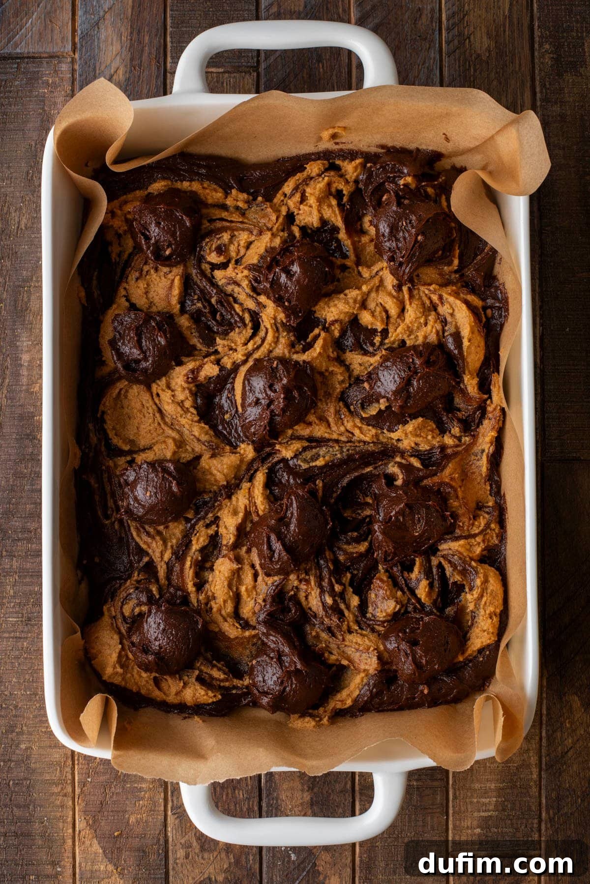 Pumpkin Ripple Fudge Brownies 10 A rectangular baking dish lined with parchment paper holds a freshly baked batch of chocolate and pumpkin swirled brownies, with rich dark and vibrant orange swirls beautifully marbled on top.