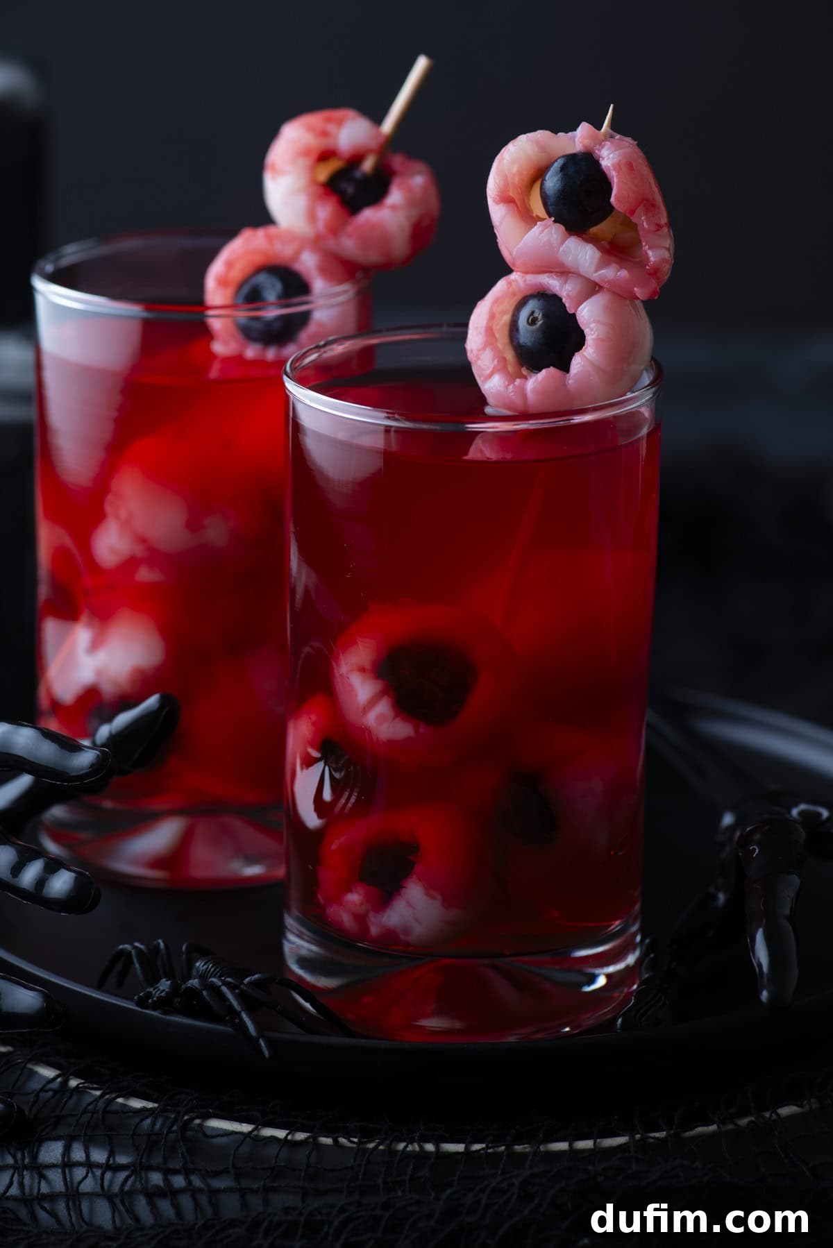 Two glasses filled with red liquid, each containing peeled lychees stuffed with blueberries to resemble eyeballs, garnished with skewered lychee eyeballs on top. A black plastic hand decorates the tray beneath.