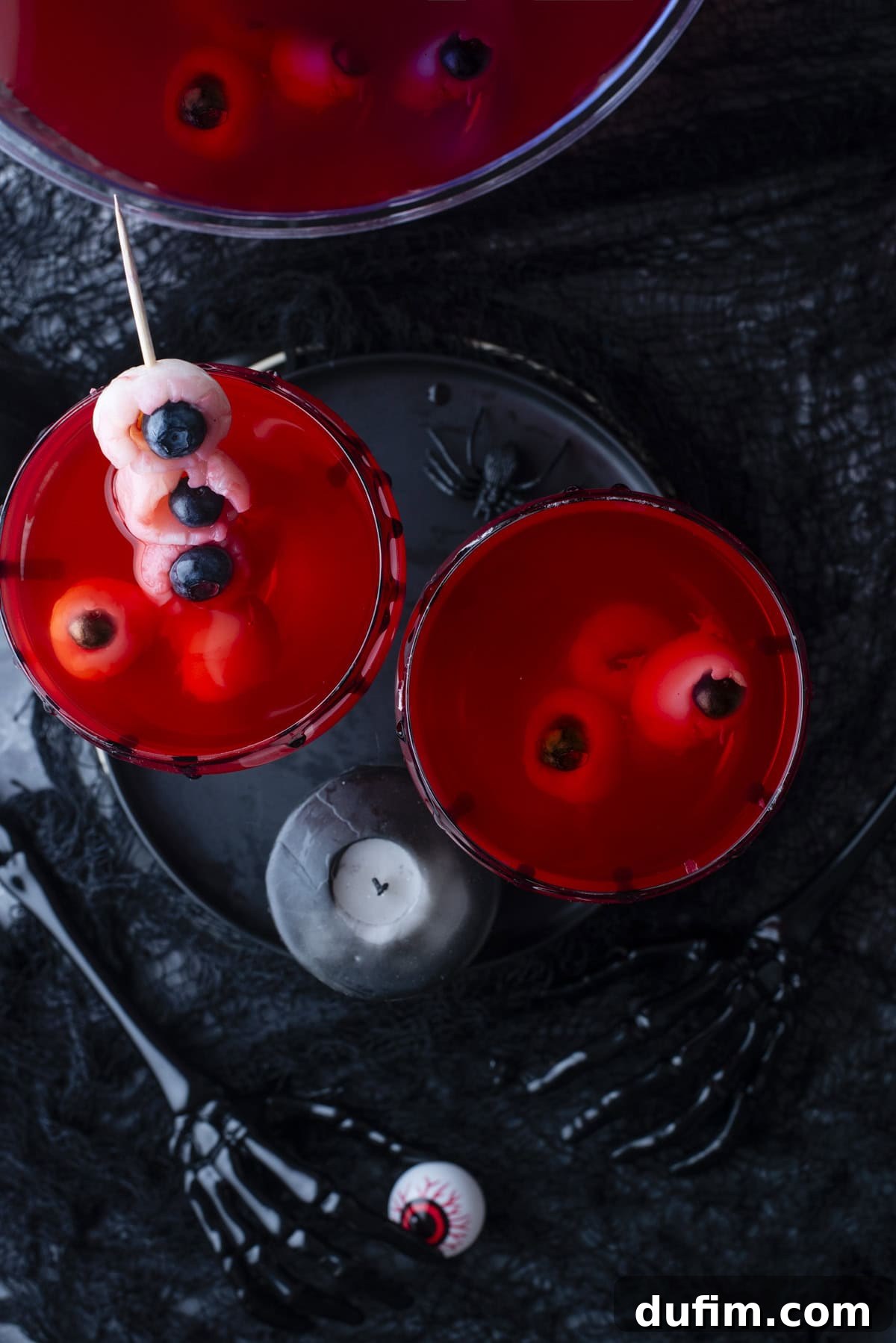 Overhead view of red drinks in glasses with eyeball-shaped garnishes, surrounded by black Halloween-themed decorations and skeleton hand utensils. A black candle and a fake eyeball are also visible.