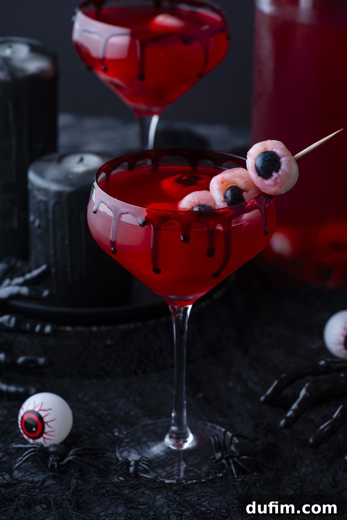 A spooky red cocktail in a glass with black drip detail, garnished with eyeball-shaped treats on a skewer. Black candles, a plastic hand, and fake eyeballs add to the Halloween theme.