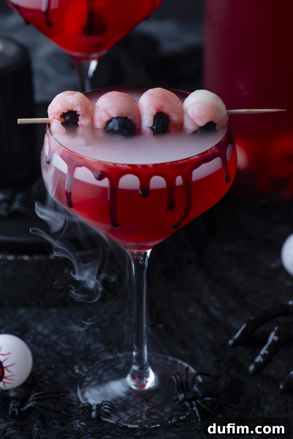 A spooky red cocktail in a glass with black syrup dripping down the rim, topped with lychee “eyeballs” on a skewer and wisps of fog, set on a dark background with Halloween decorations.