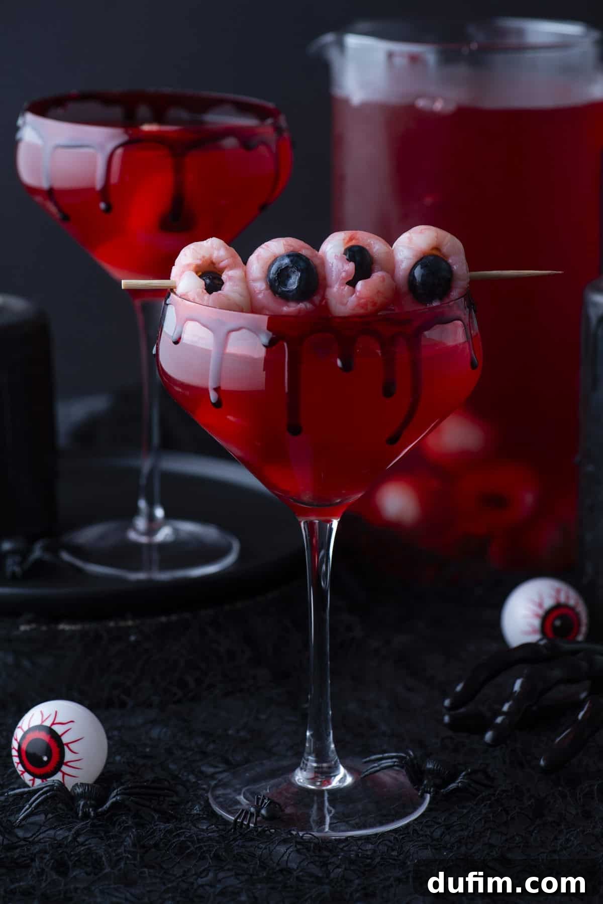 A spooky red cocktail in a glass with black syrup dripping down the rim, garnished with lychees and blueberries to look like eyeballs on a skewer. A pitcher of red drink and Halloween decorations are in the background.