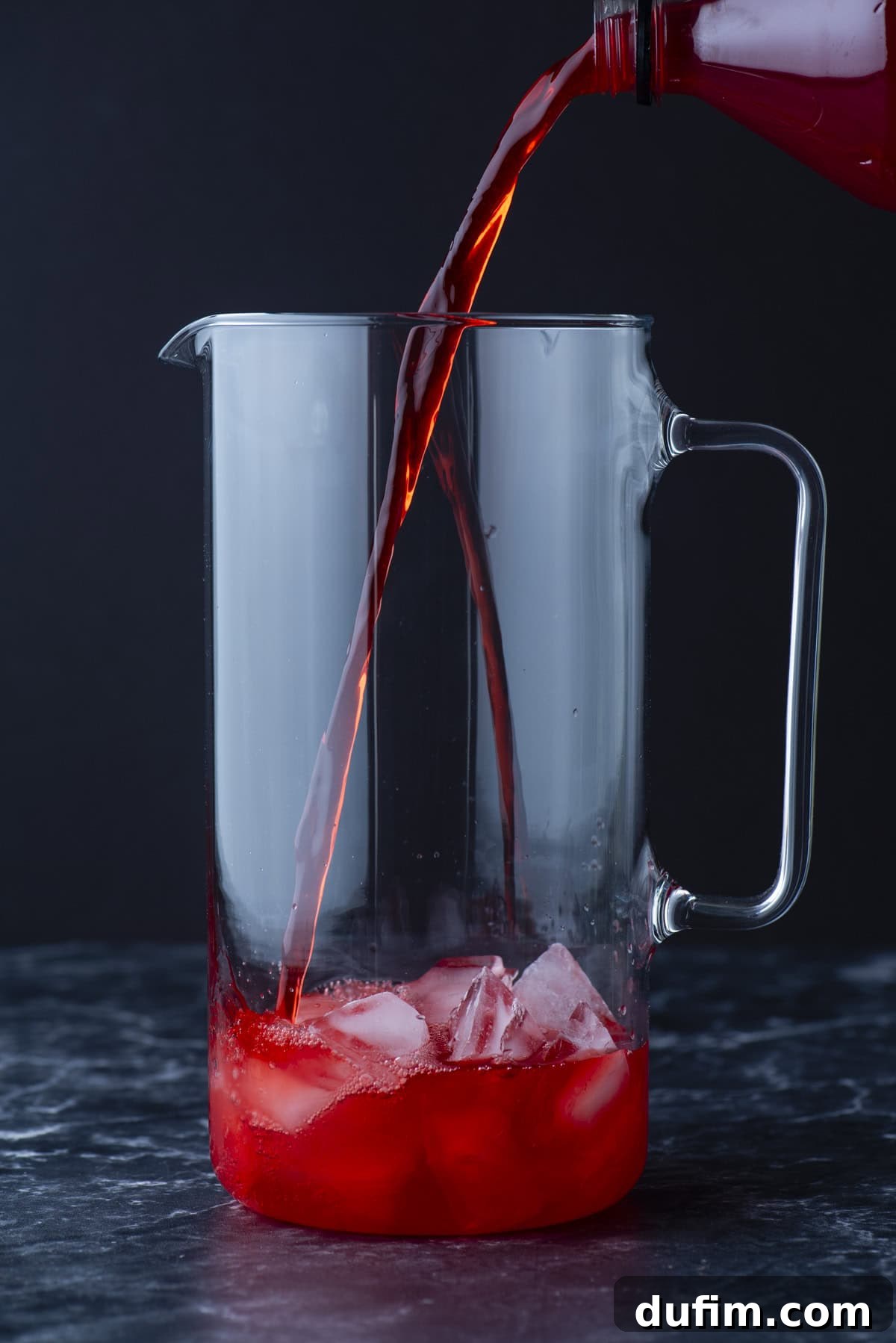 A clear glass pitcher with ice cubes inside is being filled with a red liquid poured from a bottle, set on a dark surface with a black background.