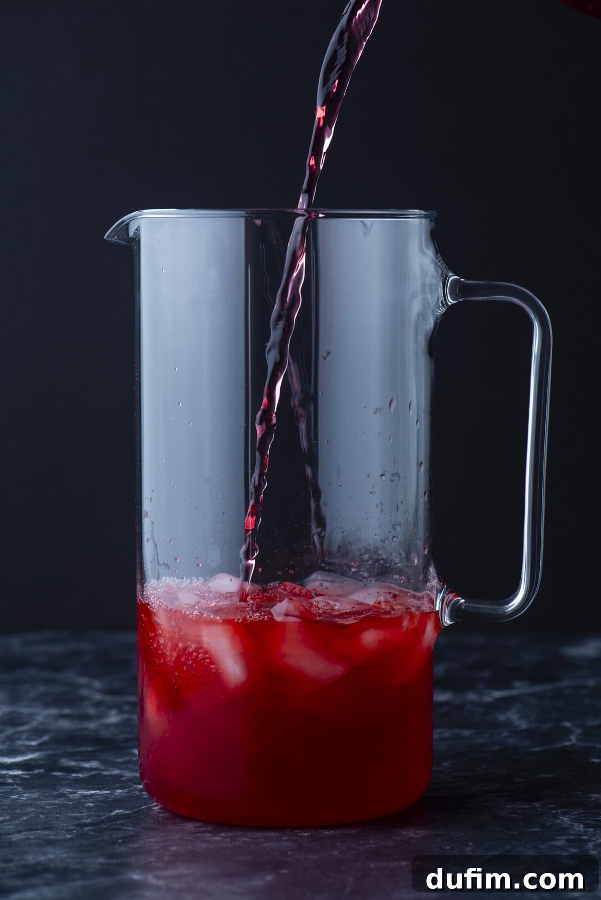 A clear glass pitcher with ice is being filled with a red liquid, possibly juice or punch, against a dark background.