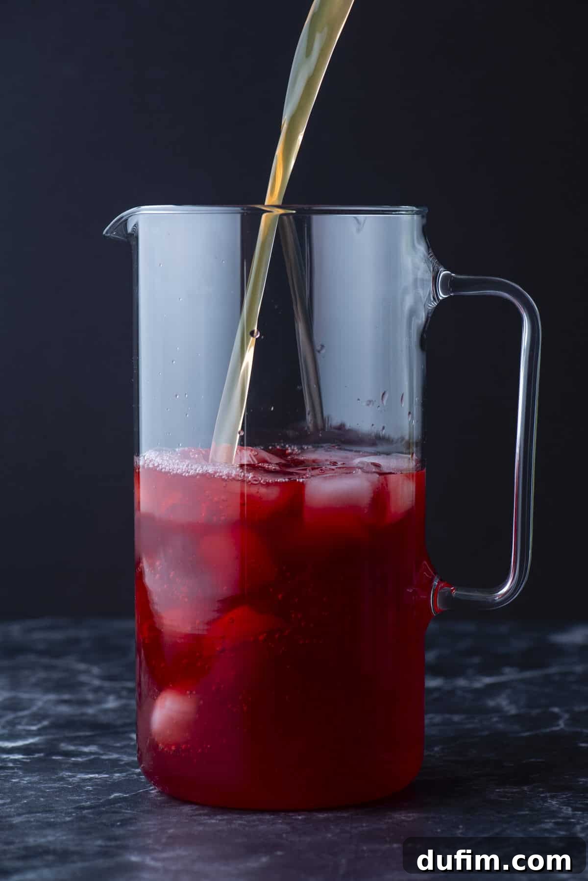 A clear glass pitcher filled with ice and red liquid, with a stream of light-colored liquid being poured in, against a dark background.