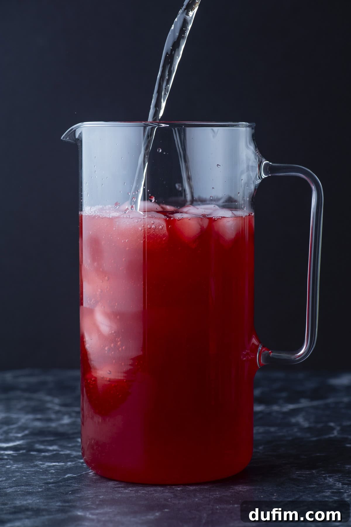 A clear glass pitcher filled with red liquid and ice, with a stream of light-colored liquid being poured in, against a dark background.