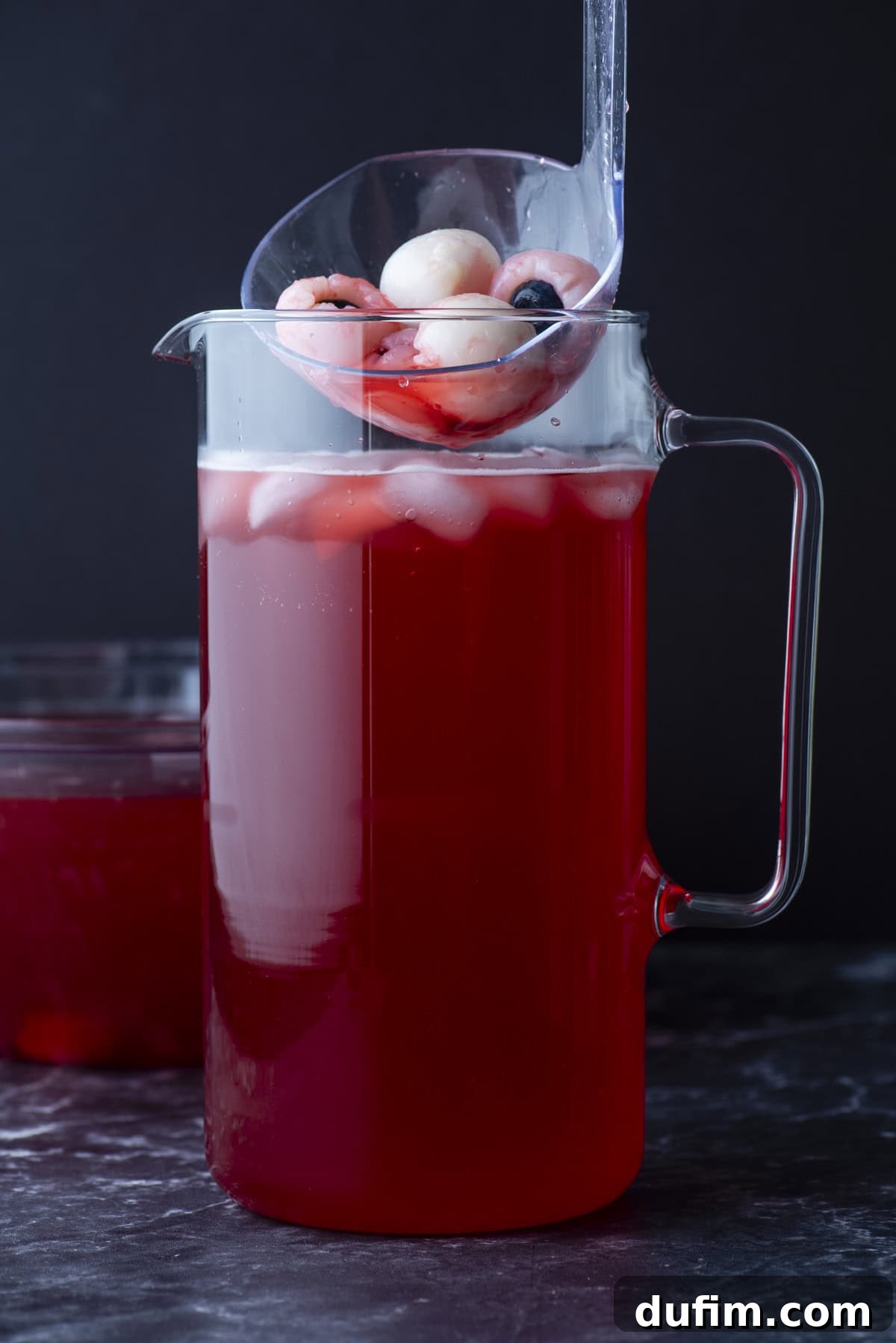 A glass pitcher filled with red punch and ice, topped with peeled lychees and blueberries. A clear ladle holds more lychees and blueberries above the pitcher. The background is dark and minimal.