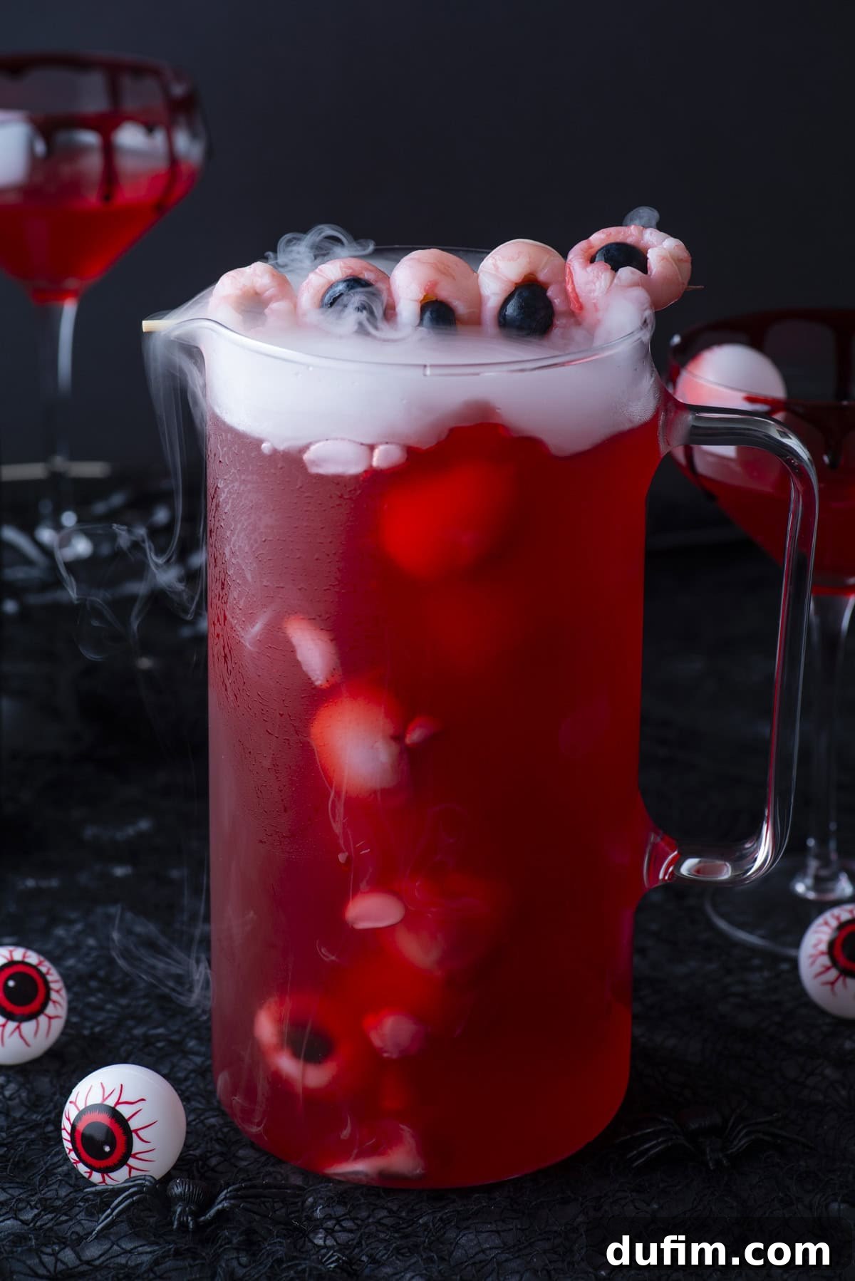 A glass pitcher of red punch with floating lychee eyeballs, blueberries, and dry ice, surrounded by plastic eyeballs and cocktail glasses, creating a spooky Halloween-themed drink scene.