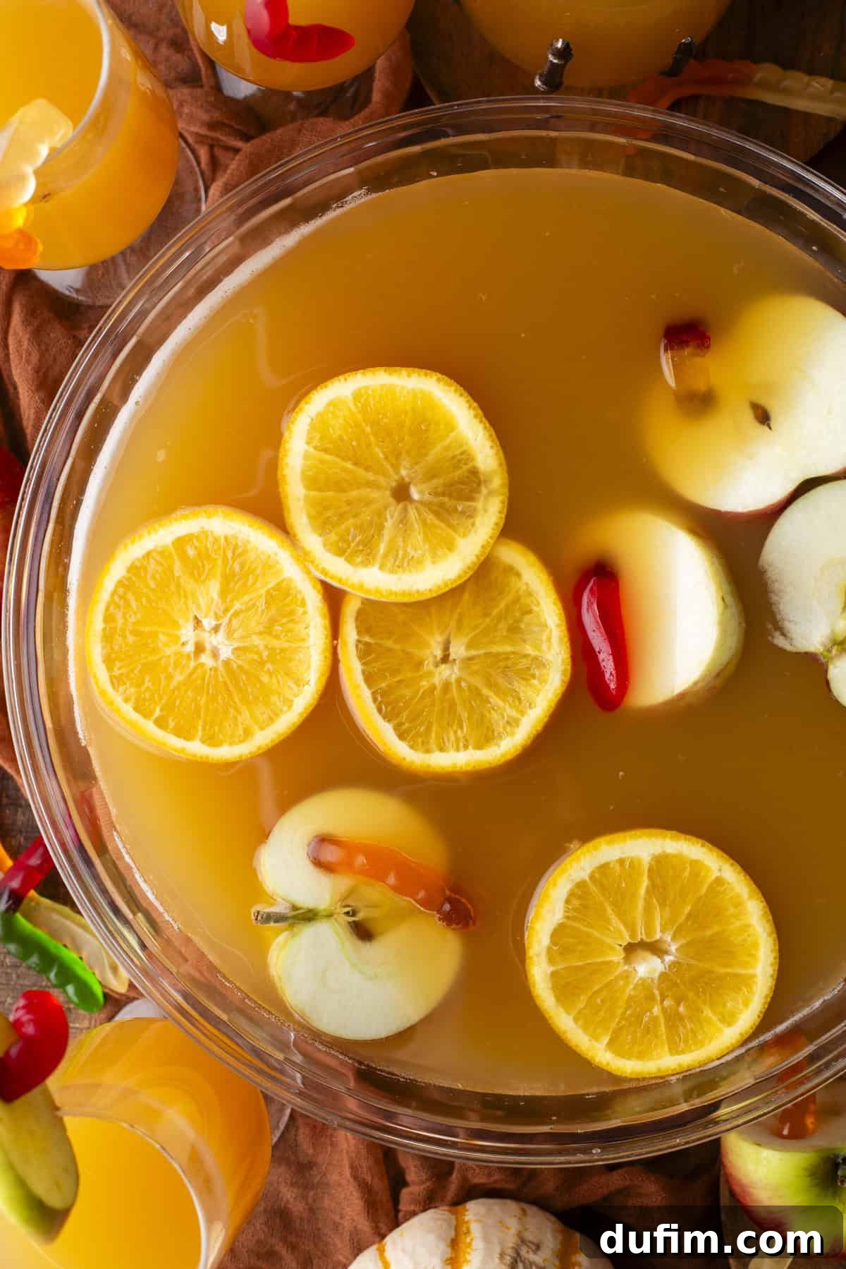 A vibrant orange-colored punch in a large bowl, garnished generously with fresh orange slices and red apple slices. Surrounding the bowl are glasses filled with the same inviting drink, nestled amongst autumn-themed decorations like small pumpkins and fall leaves, evoking a warm and festive Halloween atmosphere.