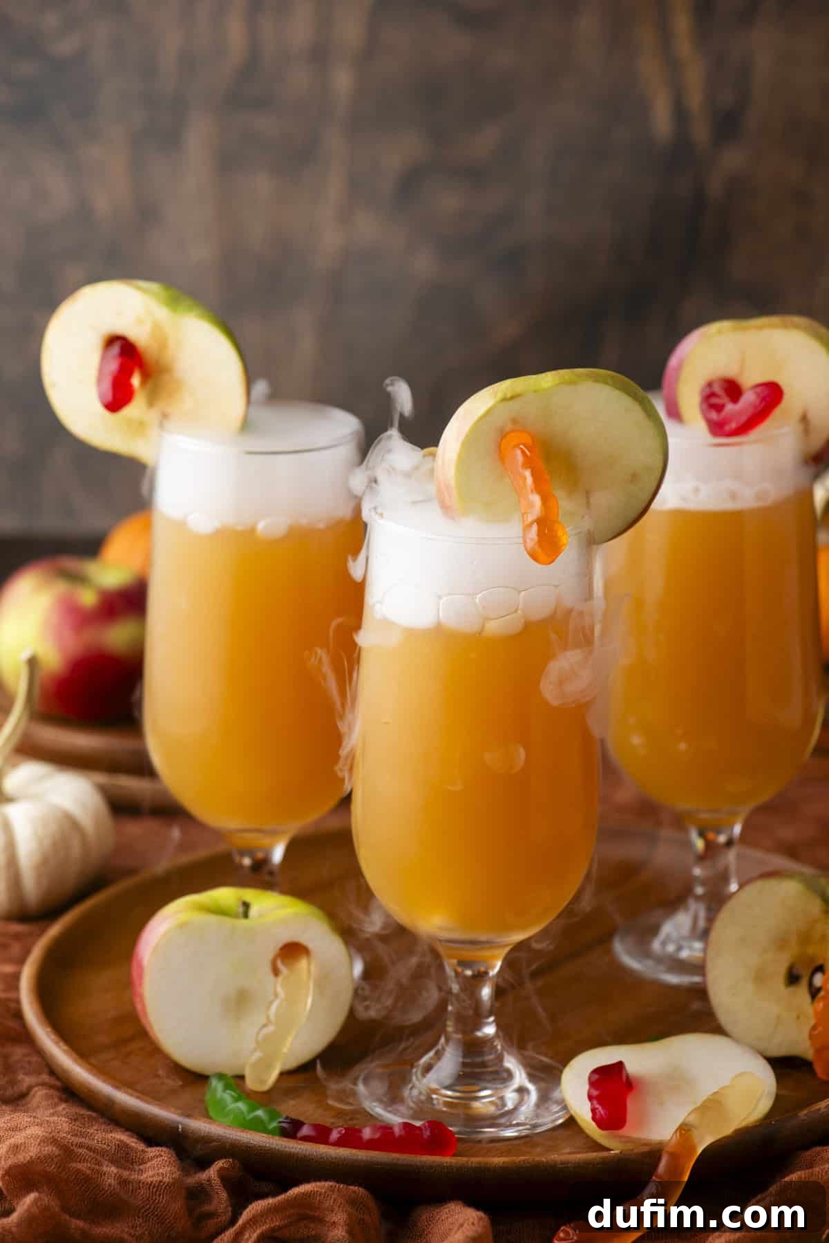 Three tall glasses containing orange Halloween cocktails, each beautifully garnished with a slice of apple and several gummy worms. Wisps of smoke or vapor emanate dramatically from the drinks, enhancing a mystical effect. The glasses are arranged on a wooden tray, surrounded by additional fresh apples and gummy candies, creating a captivating and festive autumn scene.