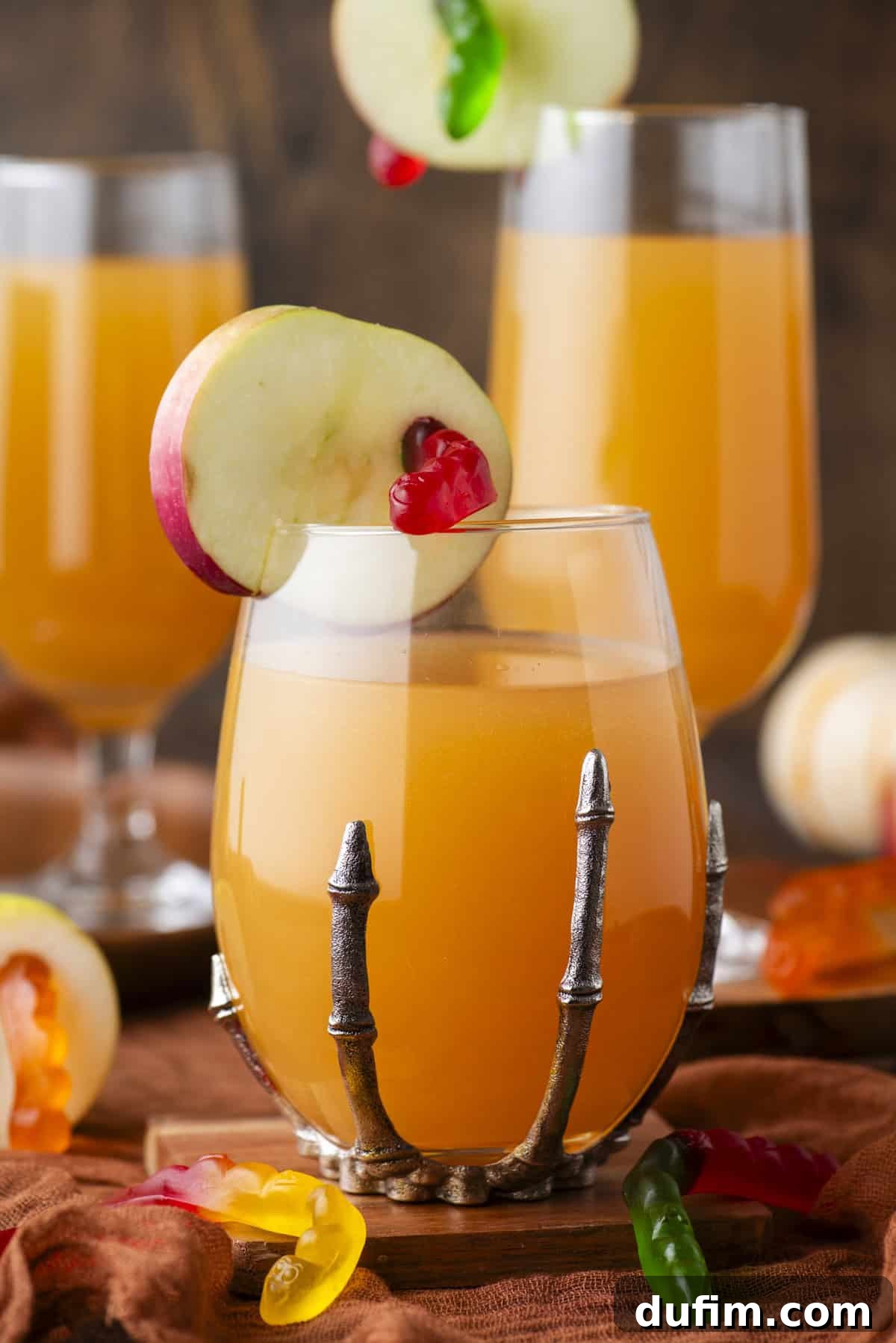 A close-up of a glass filled with a vibrant orange Halloween punch, artfully garnished with a bright red apple slice and playful gummy candies. The glass is held securely in a decorative, skeletal hand-shaped metal holder, with two similar drinks and more gummy treats visible in the softly blurred background, enhancing the spooky party theme.