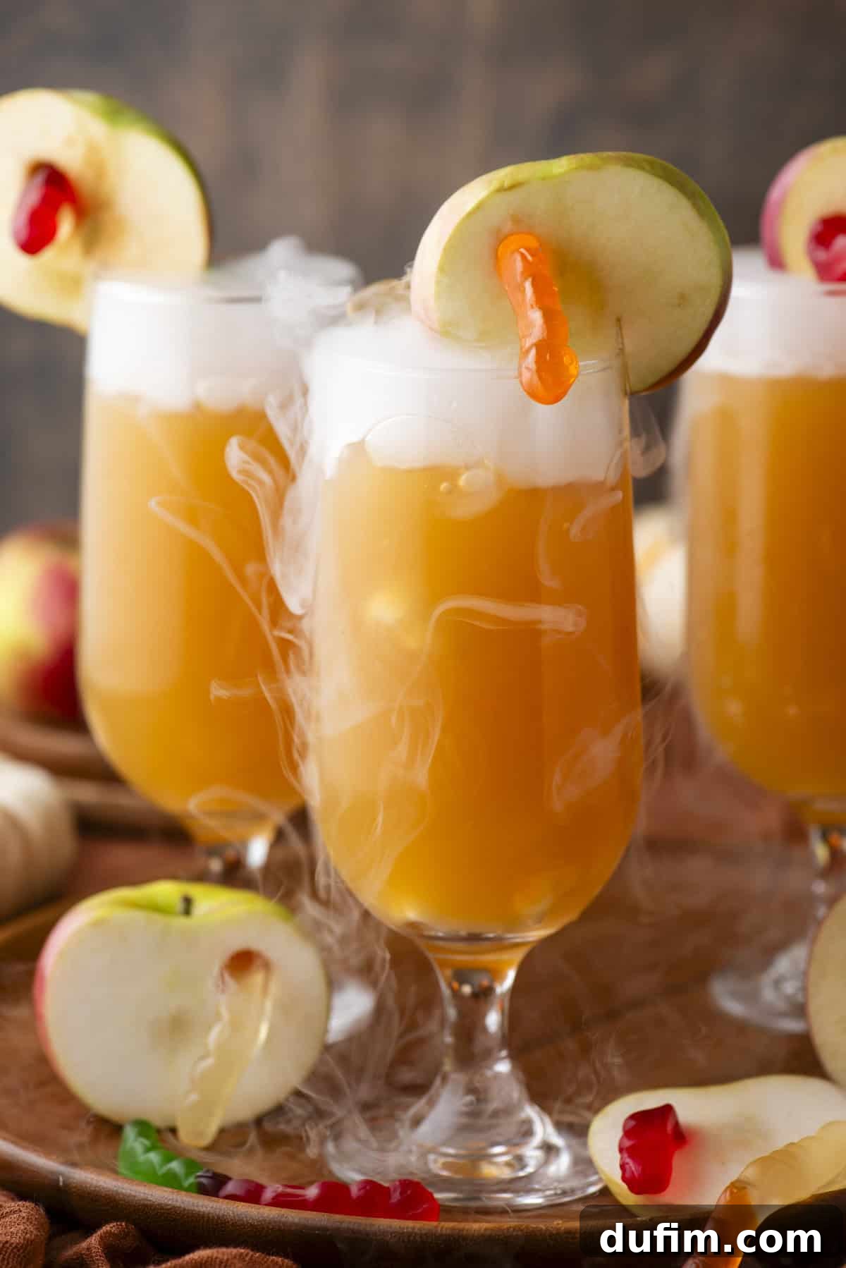 Three glasses of foamy orange punch, each garnished with an apple slice and a gummy worm, are surrounded by loose apple slices and atmospheric vapor. The setting evokes a festive and playful Halloween party ambiance.