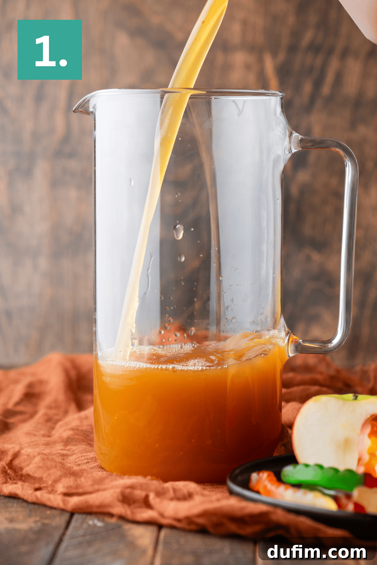 A glass pitcher is being filled with a bright orange-colored juice. The pitcher is placed on an orange fabric, with a plate of sliced apples and gummy worms visible nearby. A green box with the number 1 indicates the first step.