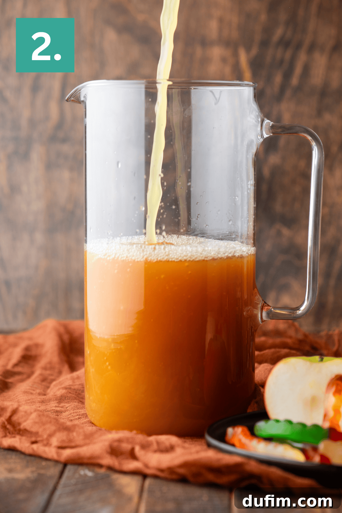 A glass pitcher is being filled with a darker orange-brown juice, indicating the addition of another ingredient. The pitcher rests on a brown cloth, next to a plate featuring an apple and gummy worms. A green box with the number 2 denotes the second step in the process.