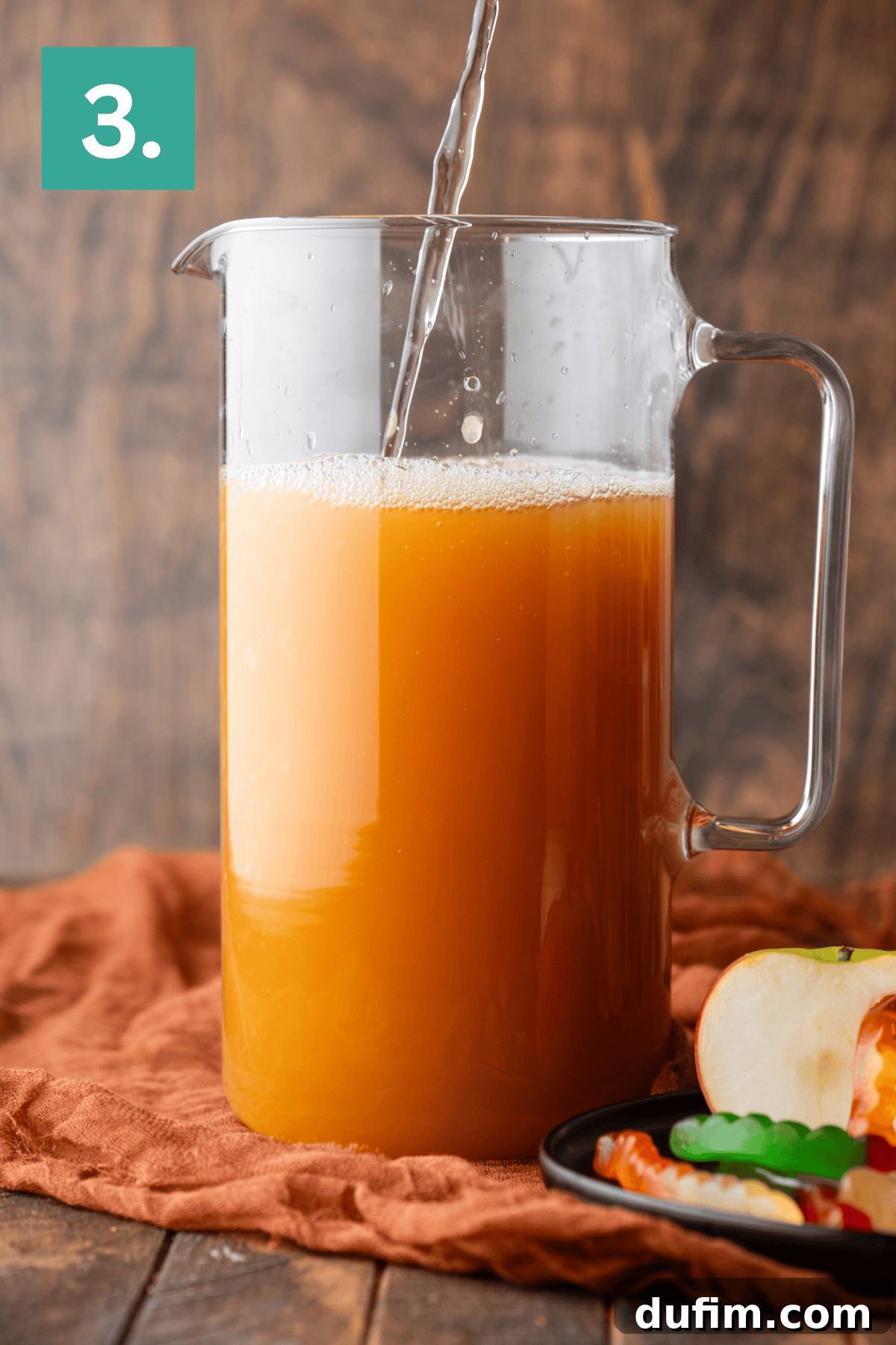 Step 3: A glass pitcher, now completely filled with the mixed orange juice, sits on a brown cloth. Liquid is still being poured, suggesting the final ingredient is being incorporated. A plate with freshly sliced apples and colorful gummy worms sits ready for the final decorative touches, showcasing the completion of the liquid mixture.